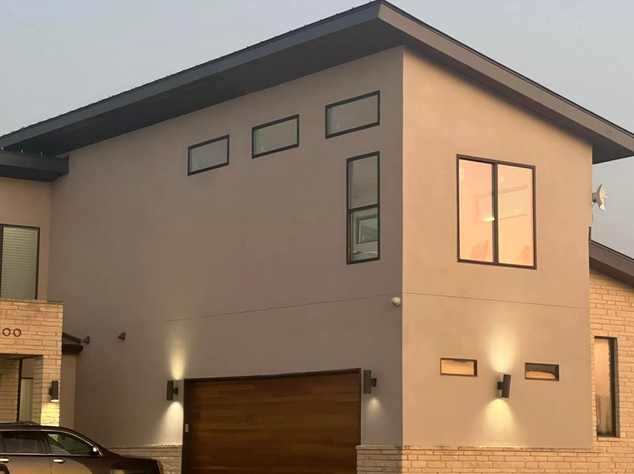 Modern two-story beige house with large windows, a garage, and exterior lights at dusk