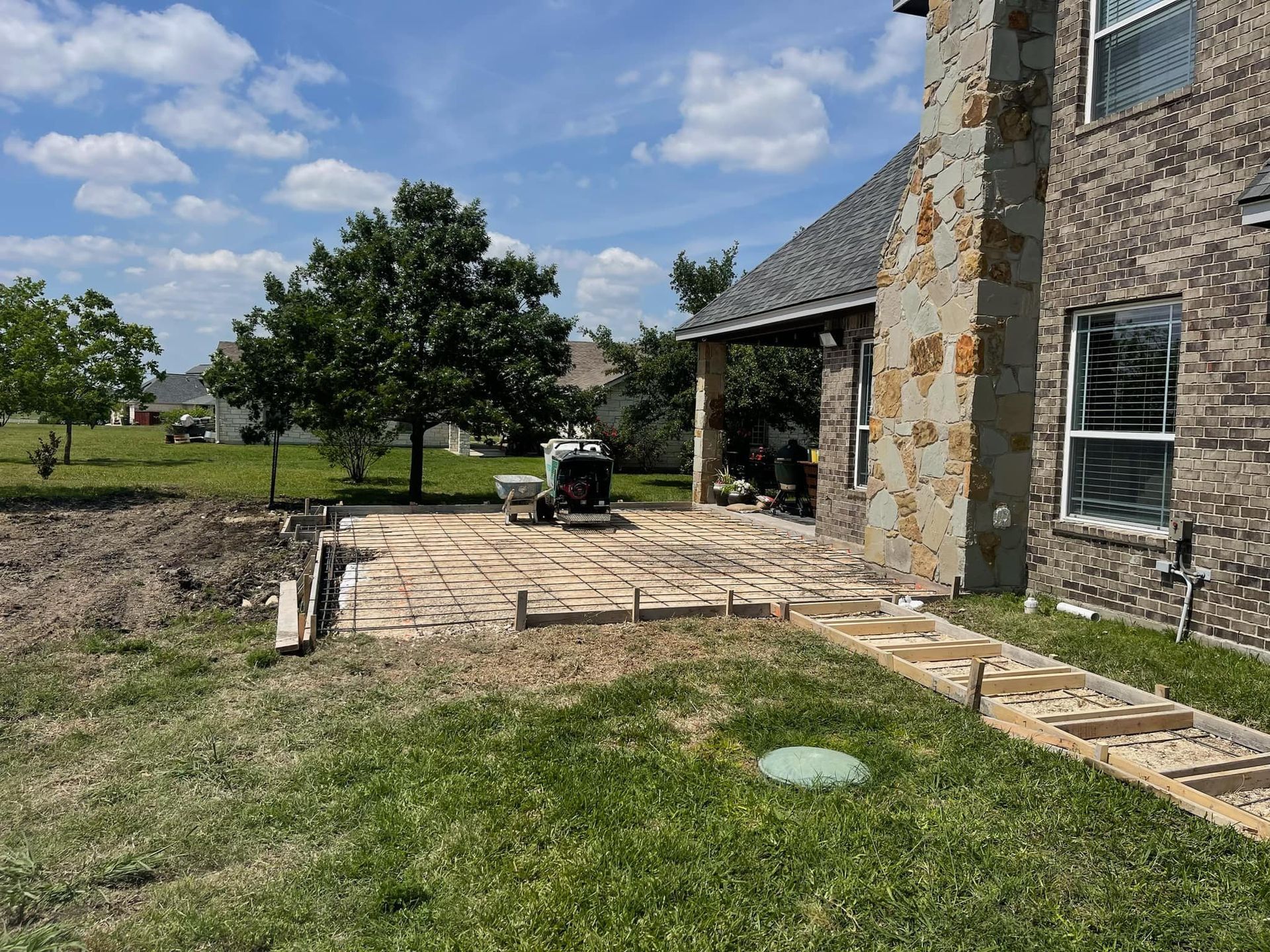 Stone patio under construction beside a brick house, with a wheelbarrow and grassy yard under a blue sky.