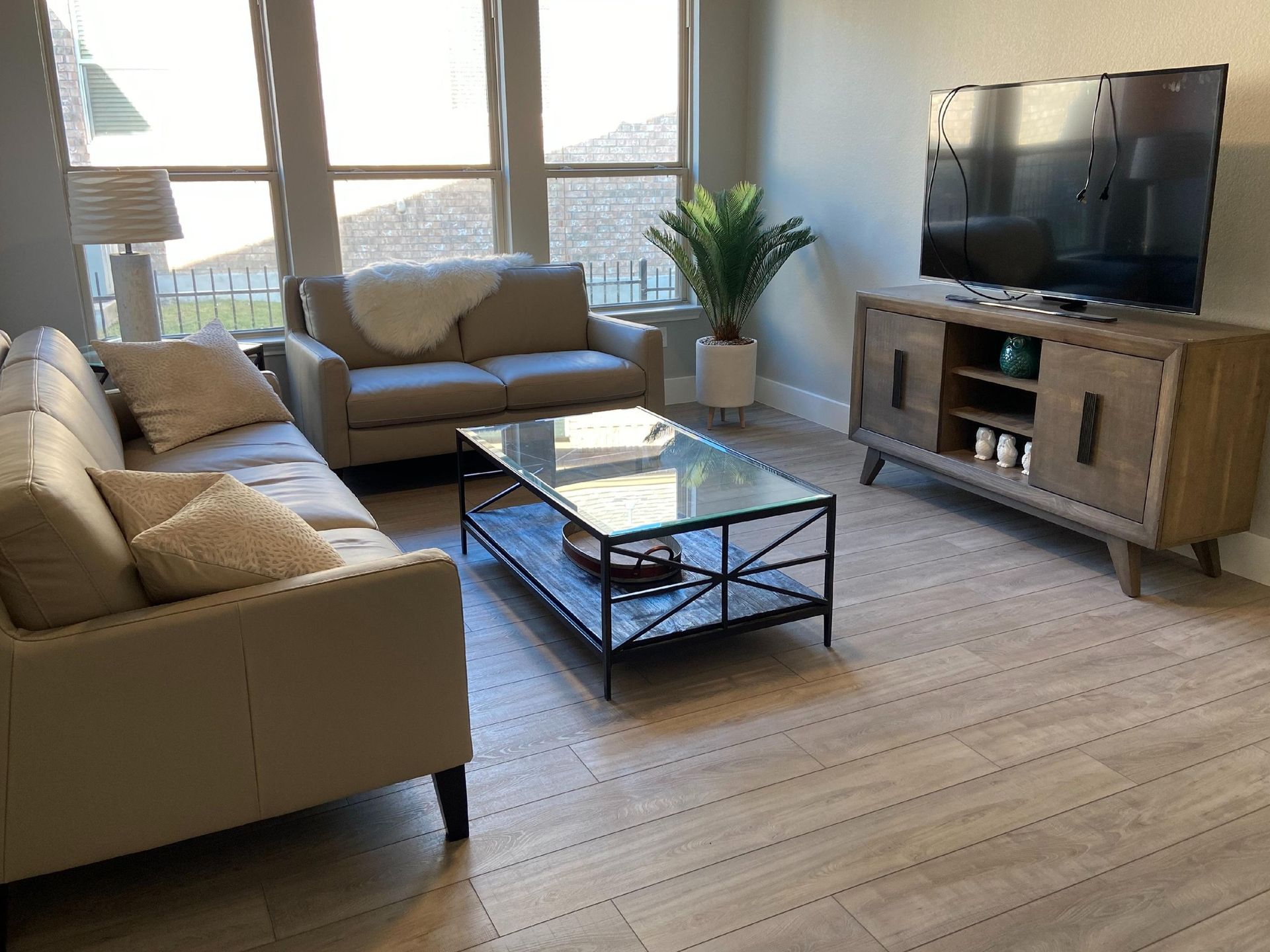 Bright living room with beige sofas, glass coffee table, TV, and large windows with sunlight