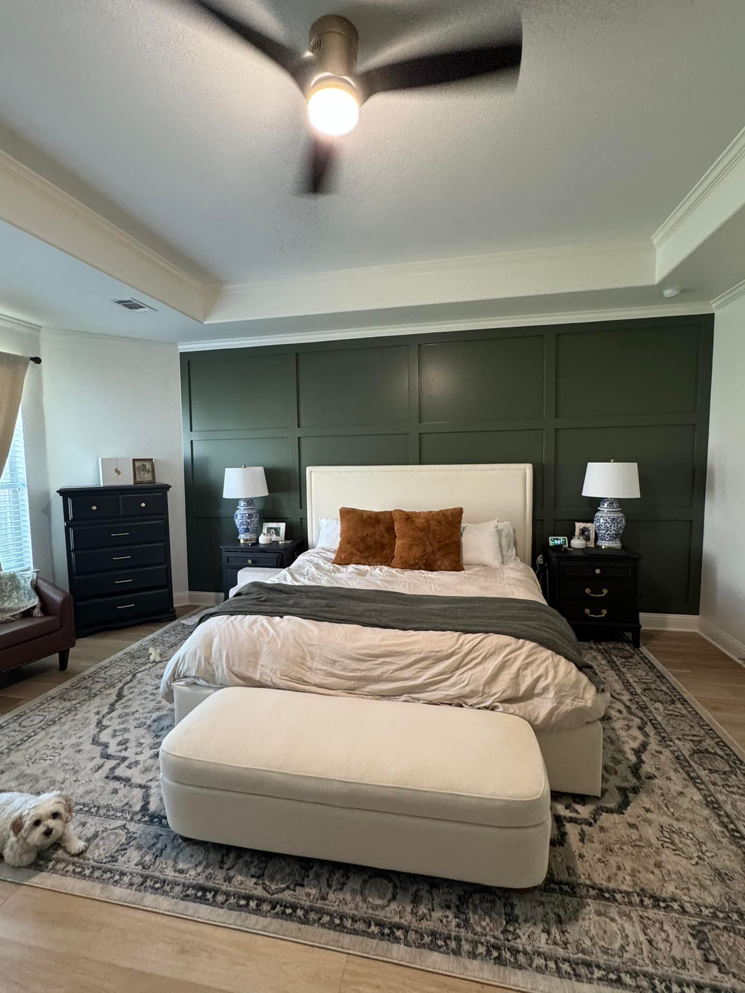 Bright bedroom with dark green wall panels, a white bed, patterned rug, and chaise under a ceiling fan.