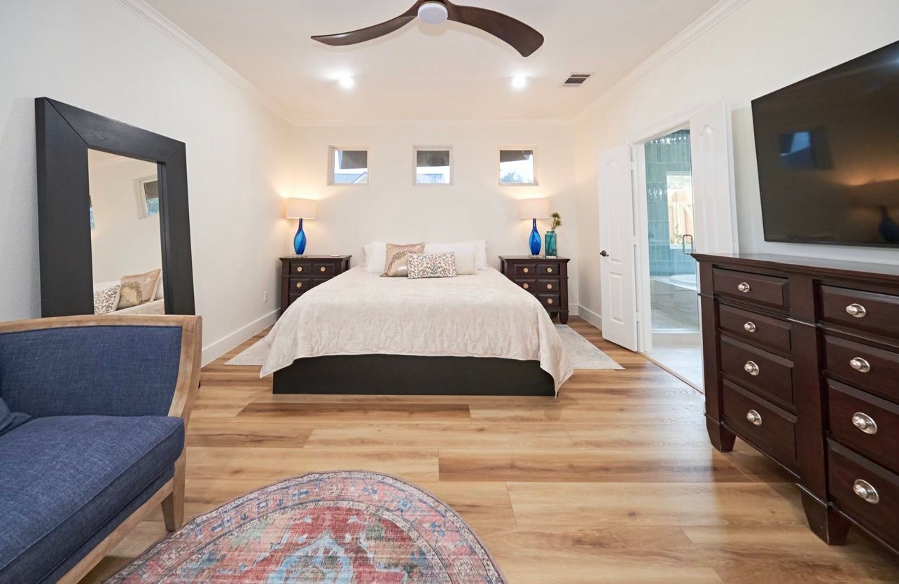 Bright bedroom with bed, blue chair, dresser, wall-mounted TV, and hardwood floors.