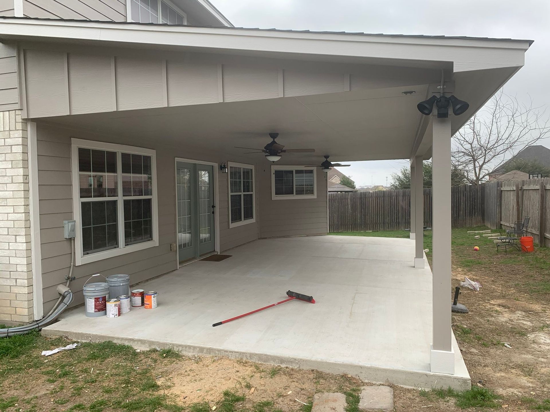 Covered concrete patio with ceiling fans, windows, and construction tools beside a brick house.