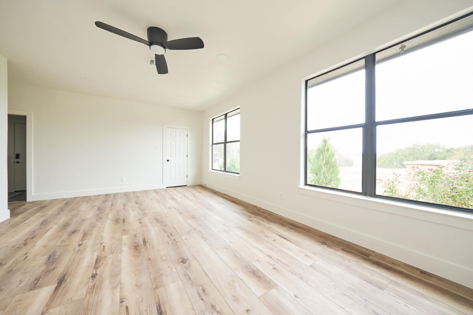 Bright empty room with light wood floors, white walls, ceiling fan, and large windows.