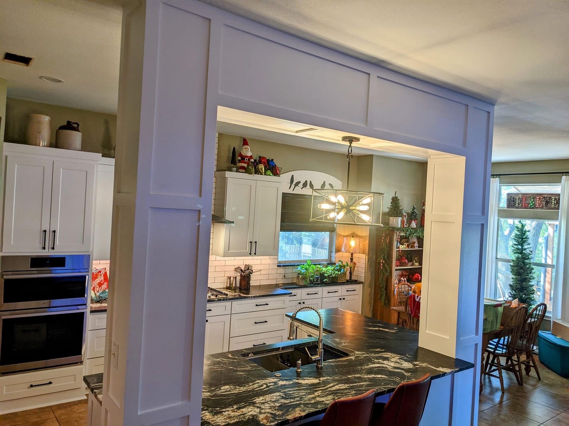 Bright kitchen with white cabinets, dark island countertop, and open view into a dining area by a window.
