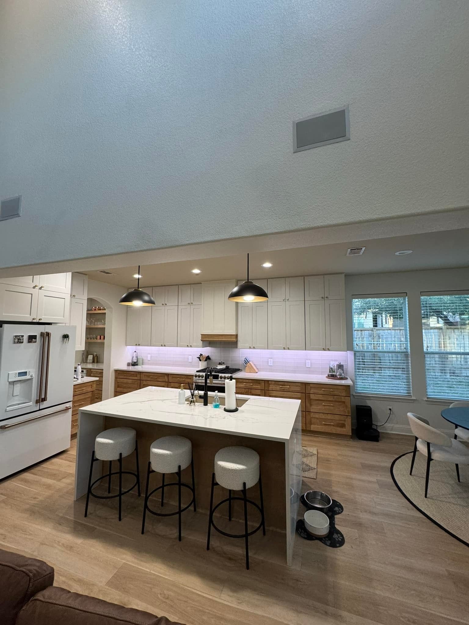 Modern kitchen with white island, hanging lights, bar stools, wood flooring, and large windows