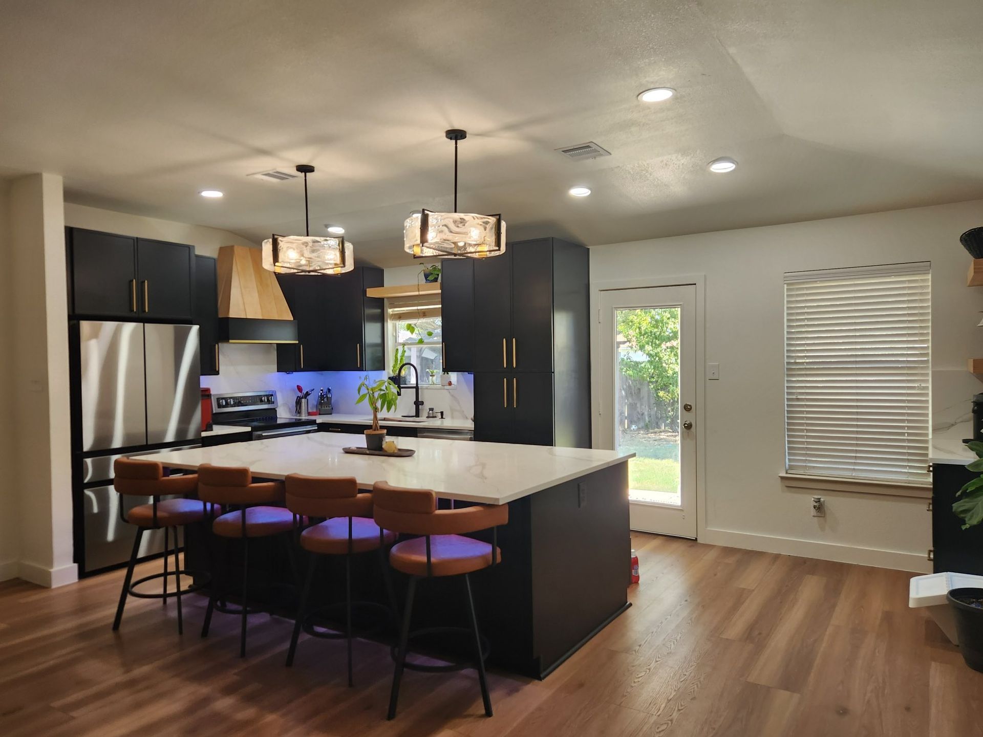Modern kitchen with island, bar stools, pendant lights, stainless fridge, and hardwood floors.