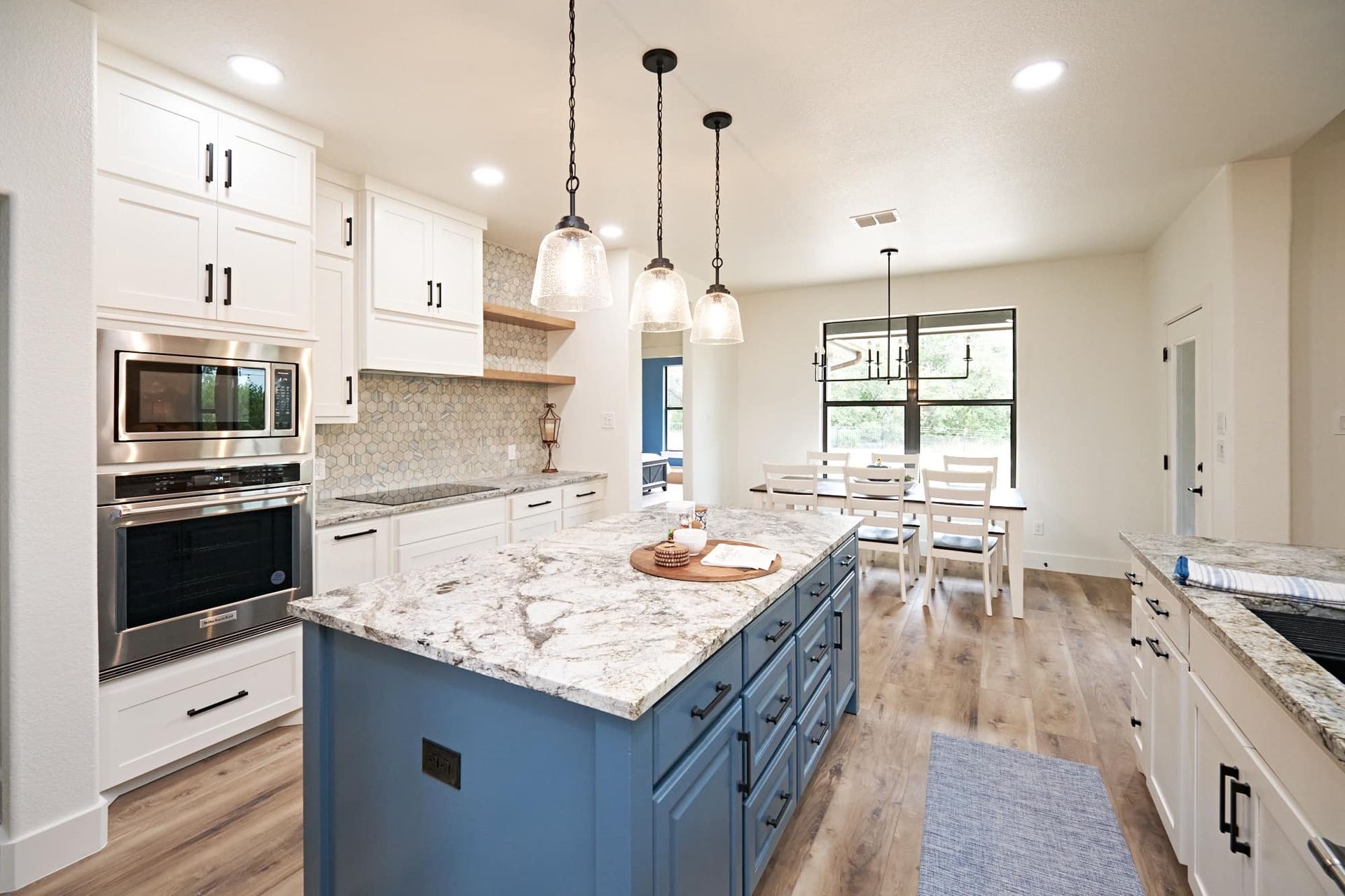 Bright modern kitchen with white cabinets, blue island, marble counters, and dining area by a window.