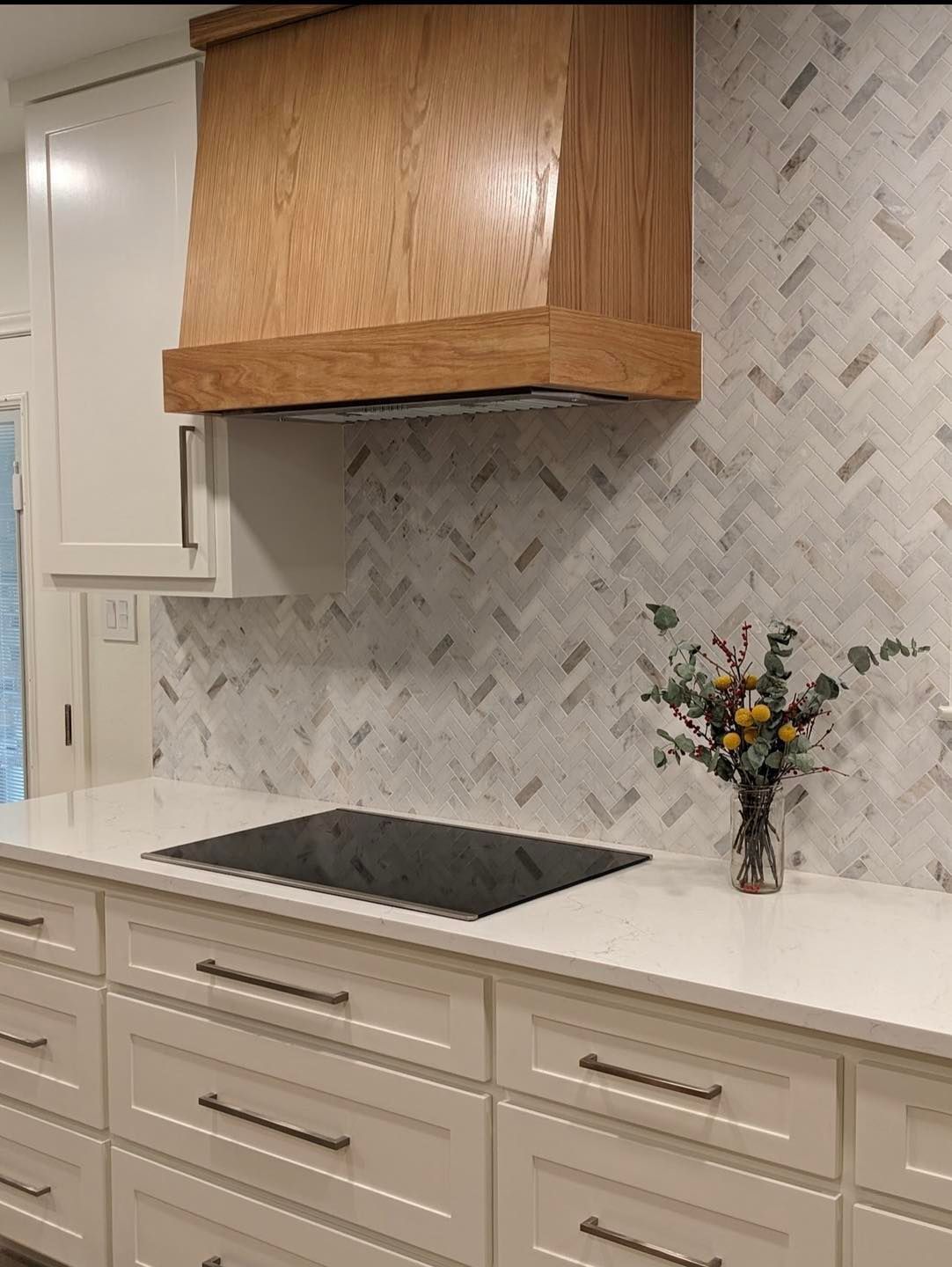 Modern kitchen with wood cabinets, white countertops, patterned backsplash, black cooktop, and small flower vase