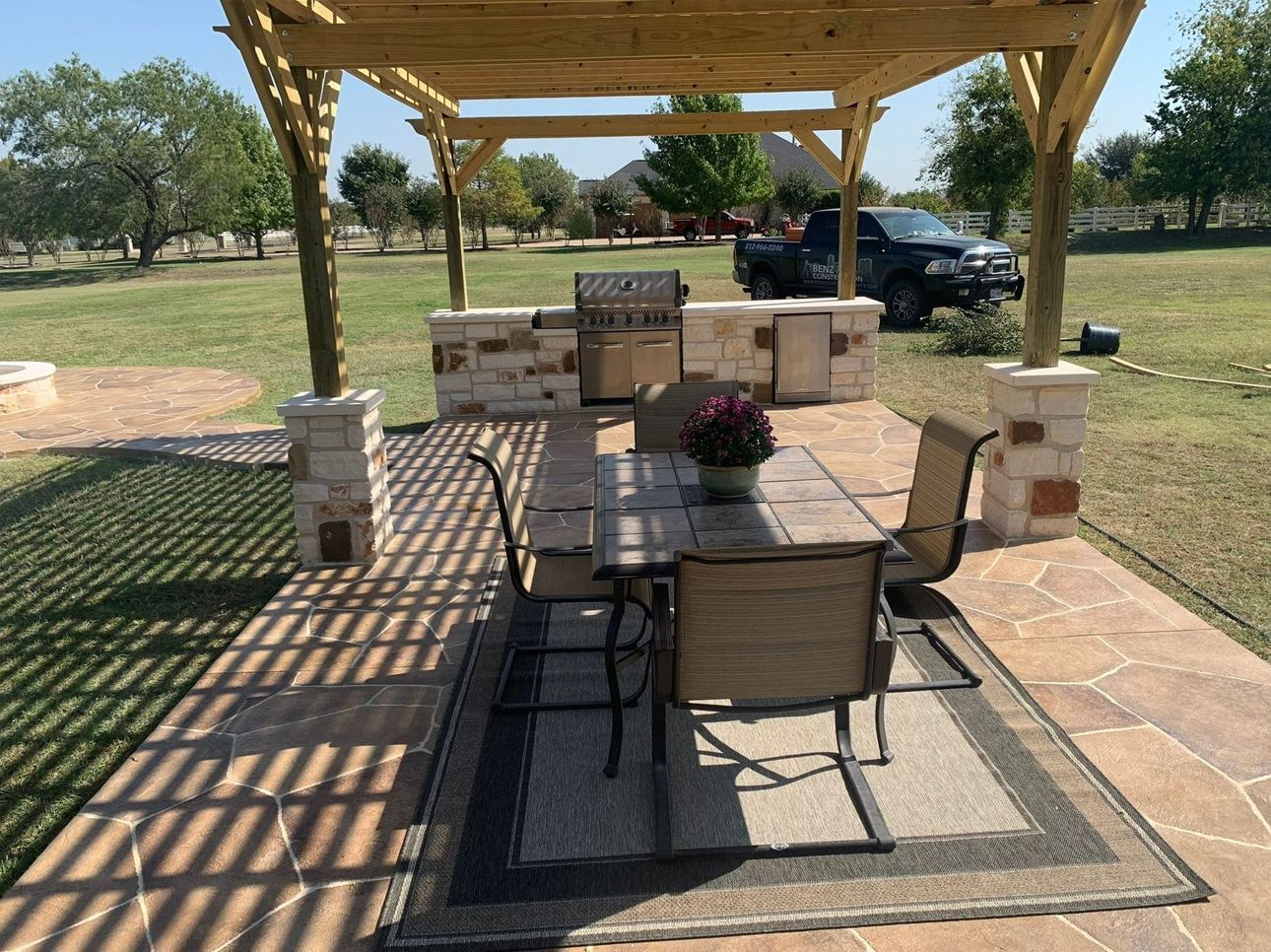 Outdoor patio with pergola, stone seating, table, and parked vehicles in a grassy yard.