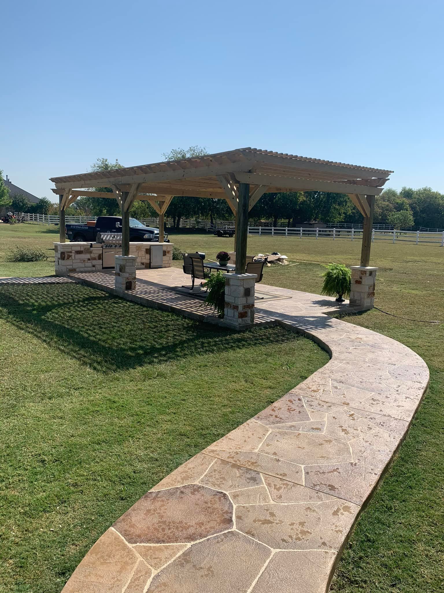 Stone path leads to a shaded picnic pavilion in a grassy park under a clear blue sky.