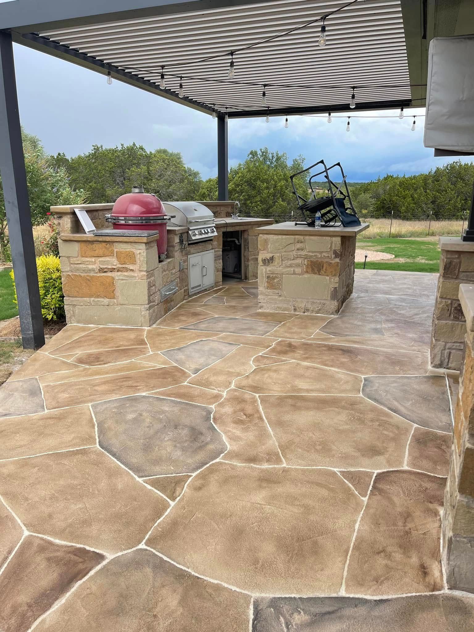 Stone patio with built-in grill and red kettle under a striped pergola, overlooking a grassy yard.