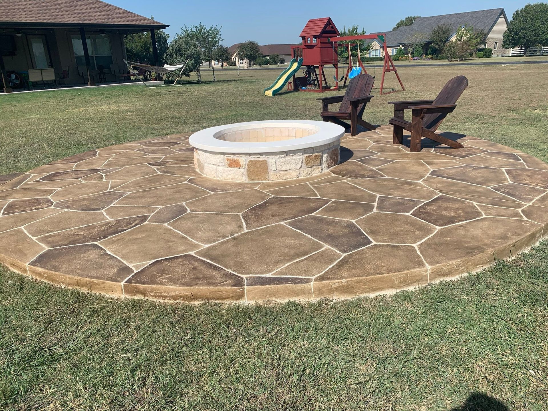 Stone fire pit centered on a circular patio with chairs, in a grassy backyard near houses and a playground.