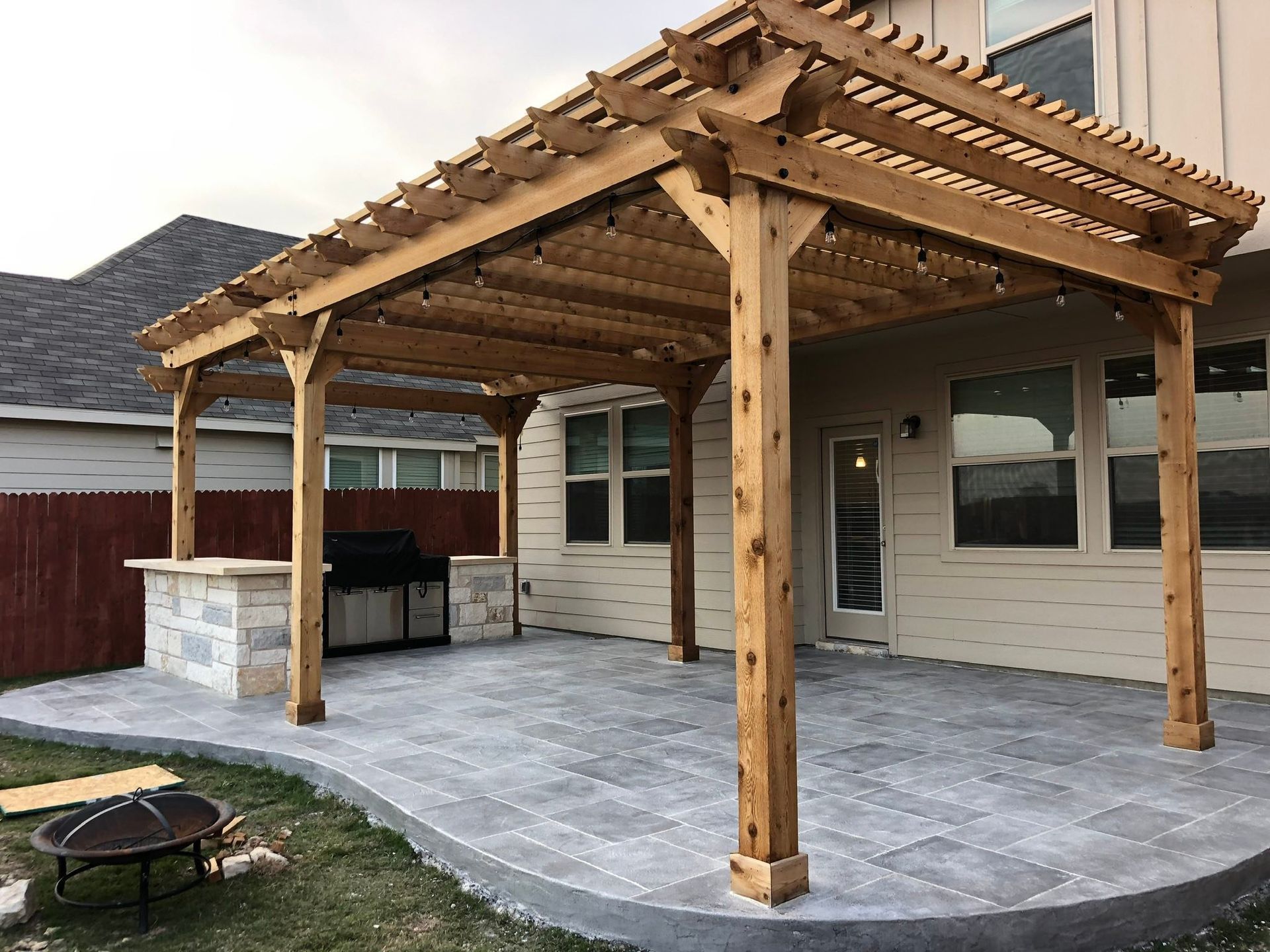 Covered wooden patio with pergola, stone flooring, and outdoor grill beside a house