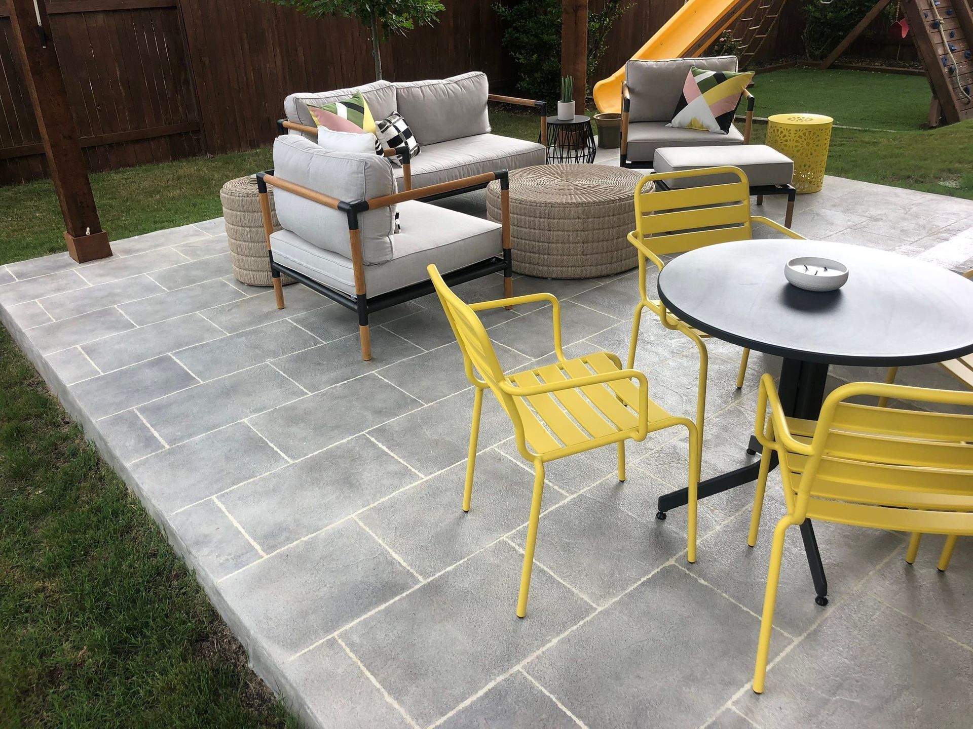 Patio with wicker sofa set, yellow chairs, round table, and gray stone paving in a backyard