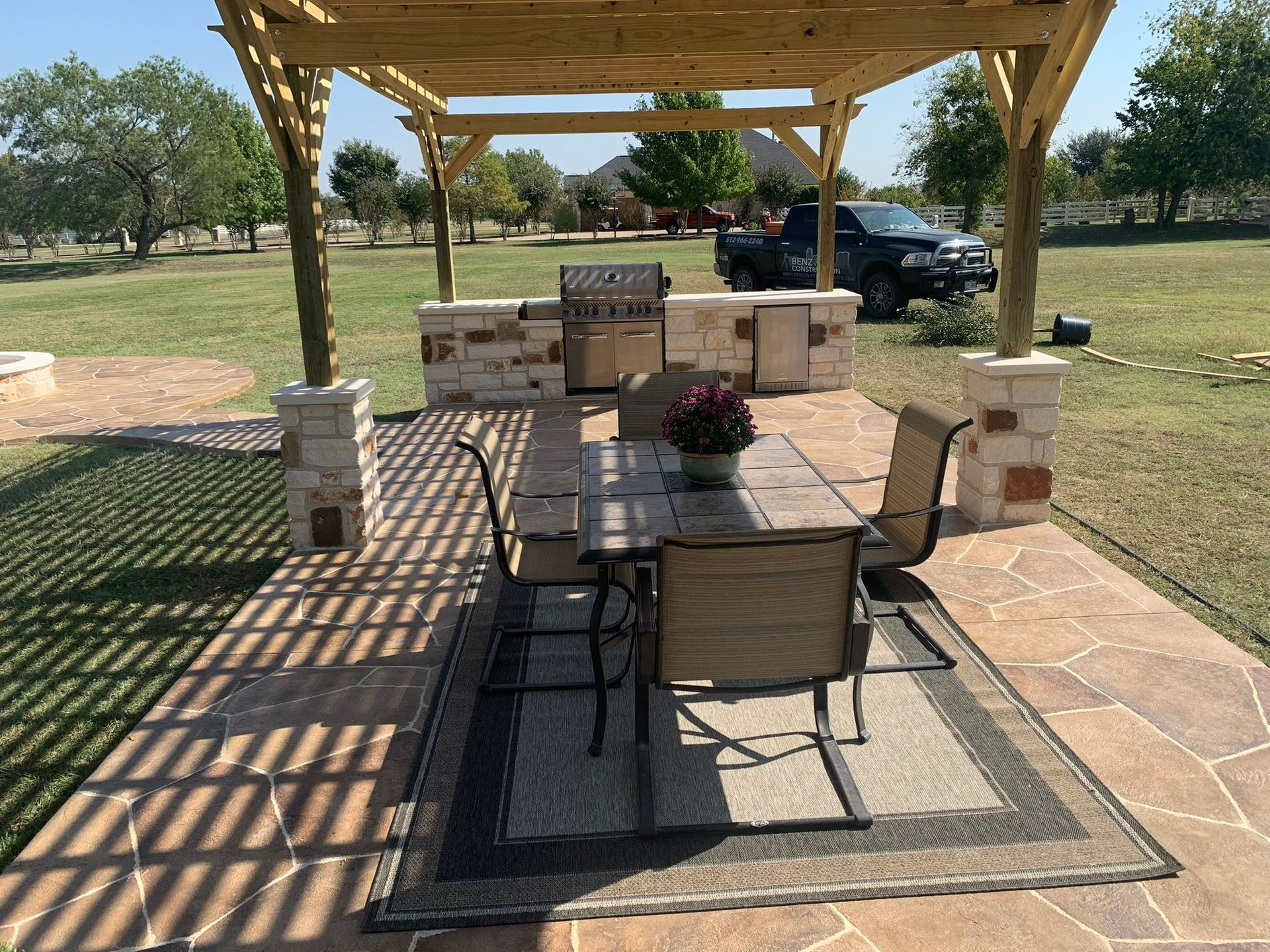 Patio dining table under a wooden pergola on a rug, with a potted centerpiece and a truck parked nearby.