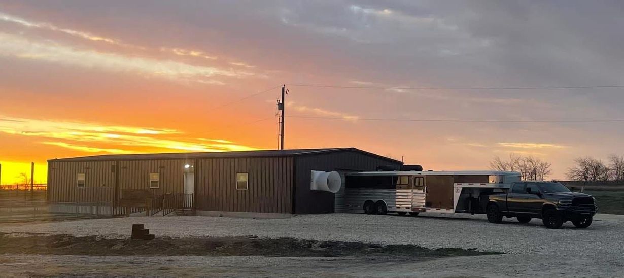 Sunset over a rural site with a long wooden shed, trucks, and equipment on a gravel lot