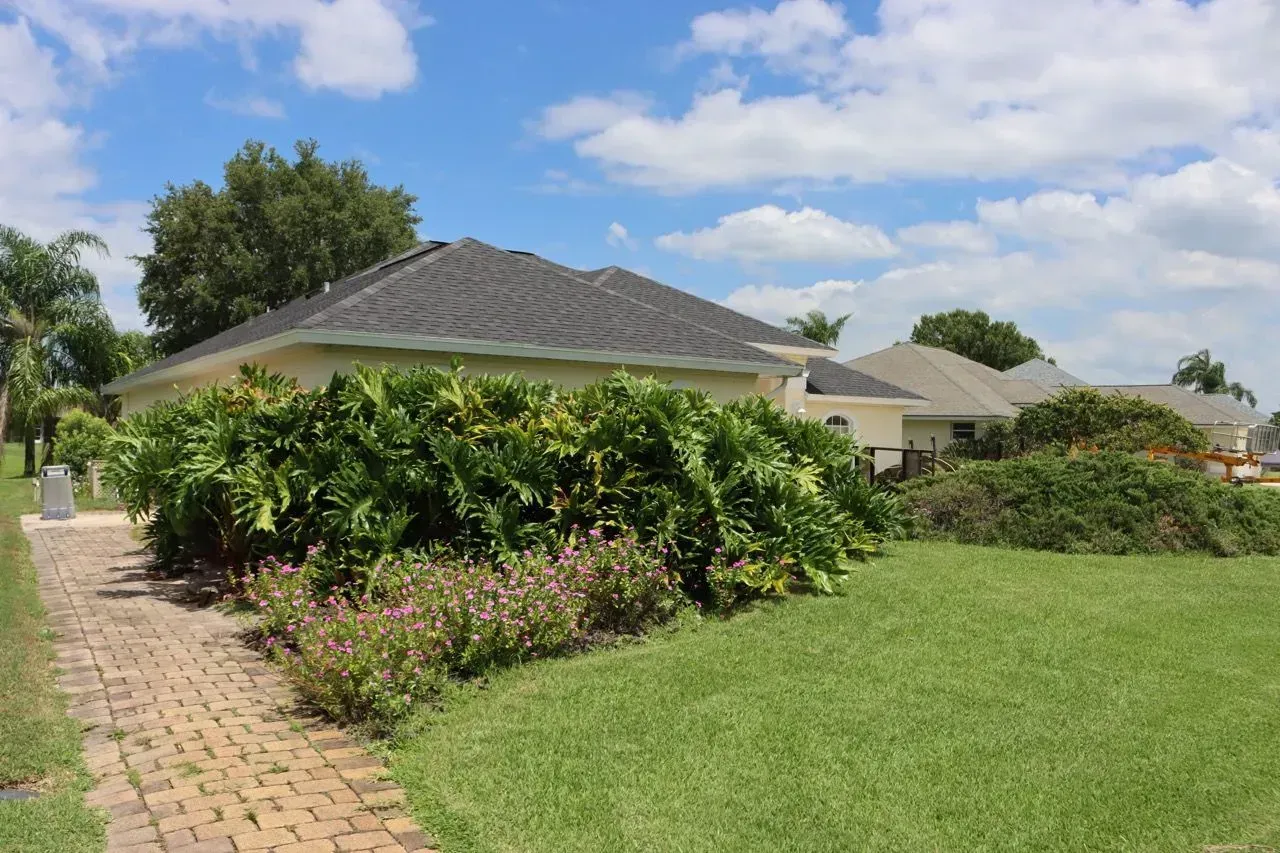 Suburban house with a tiled roof, green lawn, and bushes beside a brick walkway under a blue sky.