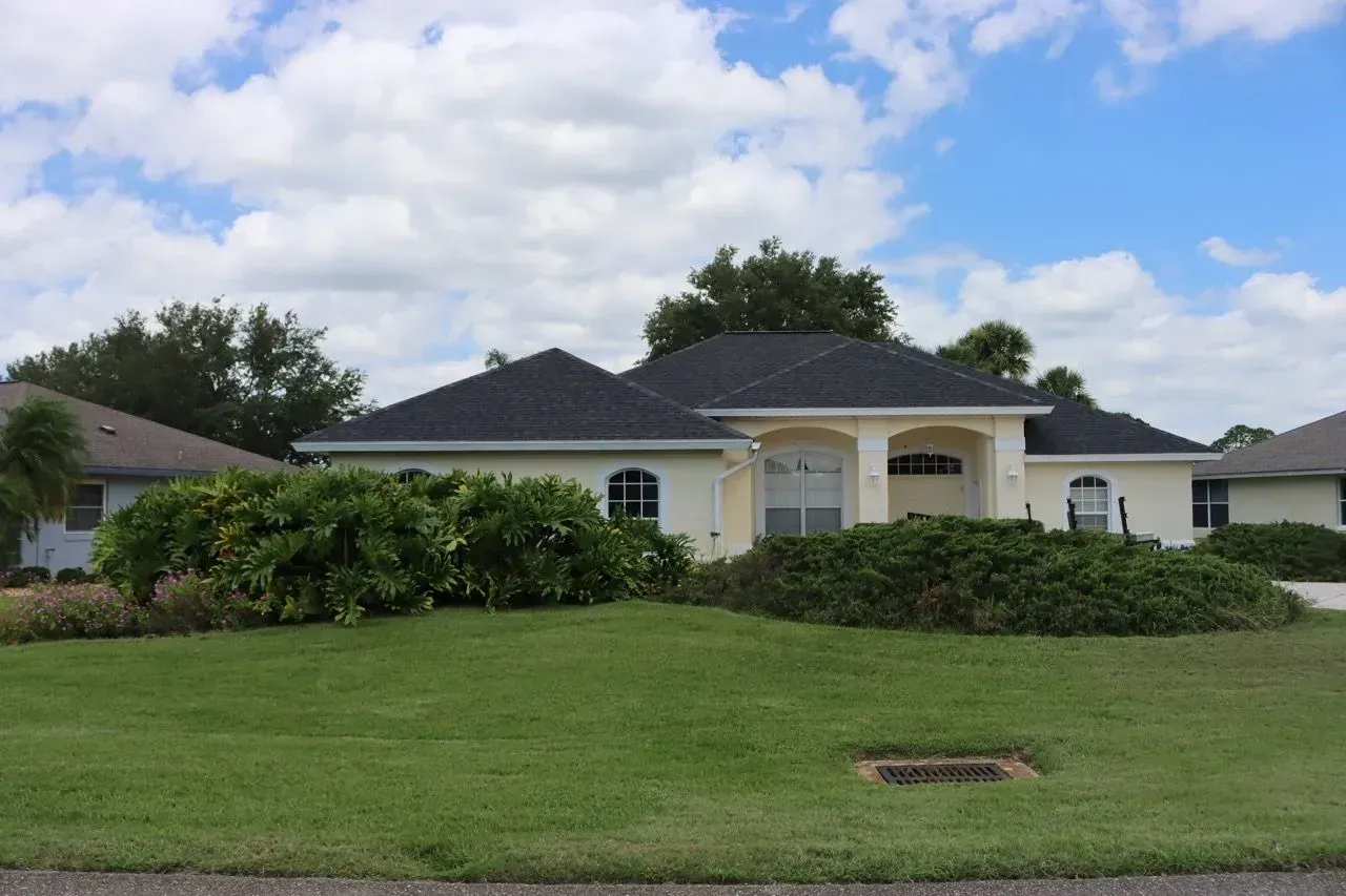 Single-story white house with dark roof, trimmed hedges, and a large green lawn under a cloudy sky