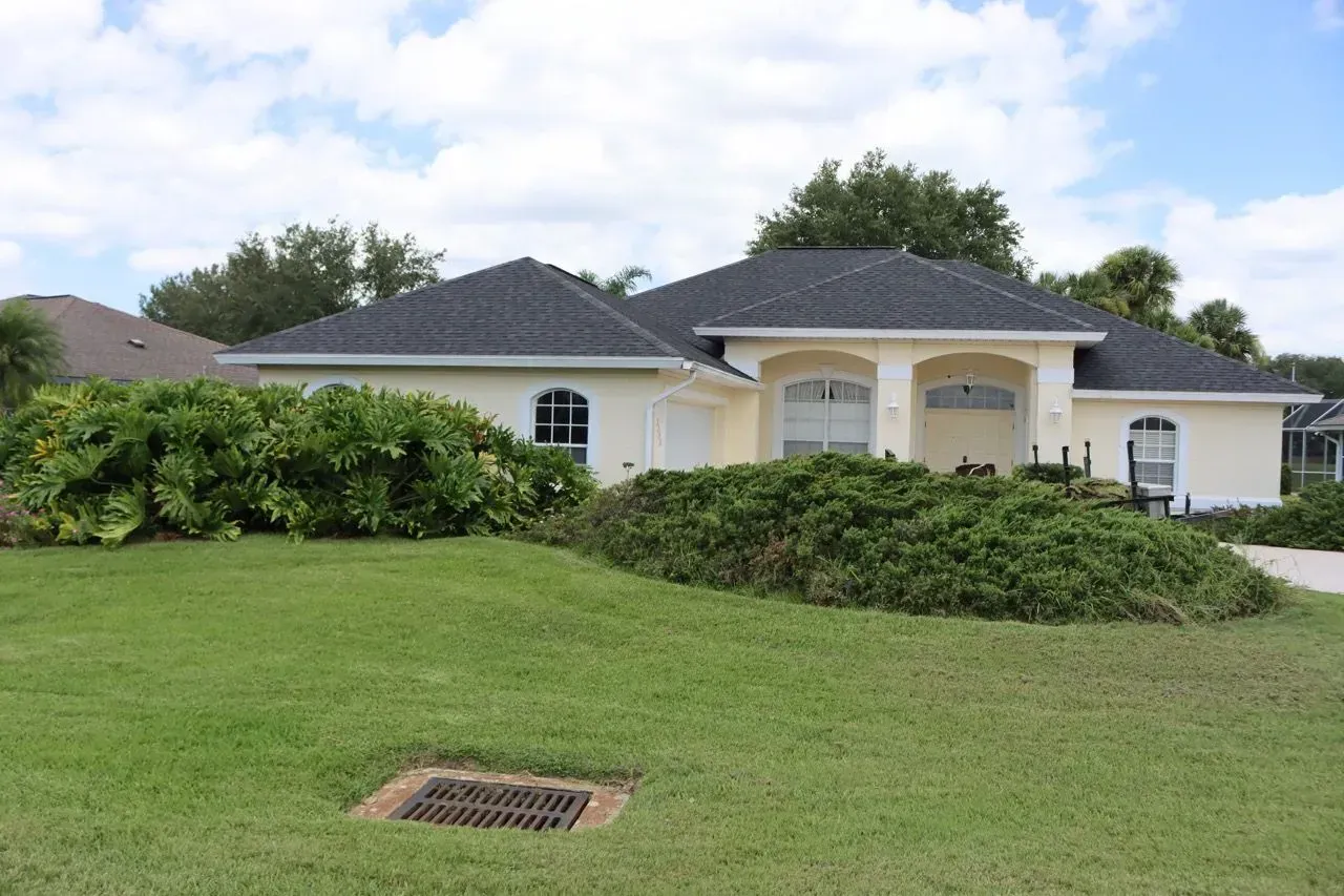 Single-story cream house with a dark roof, green lawn, shrubs, and a cloudy sky.