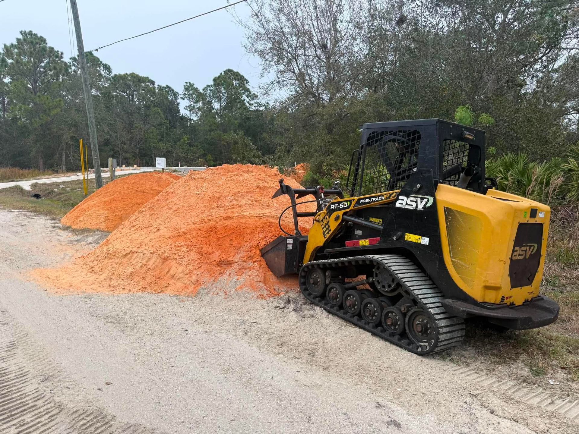 Yellow ASV track loader beside a large orange mulch pile on a gravel lot, with trees in the background
