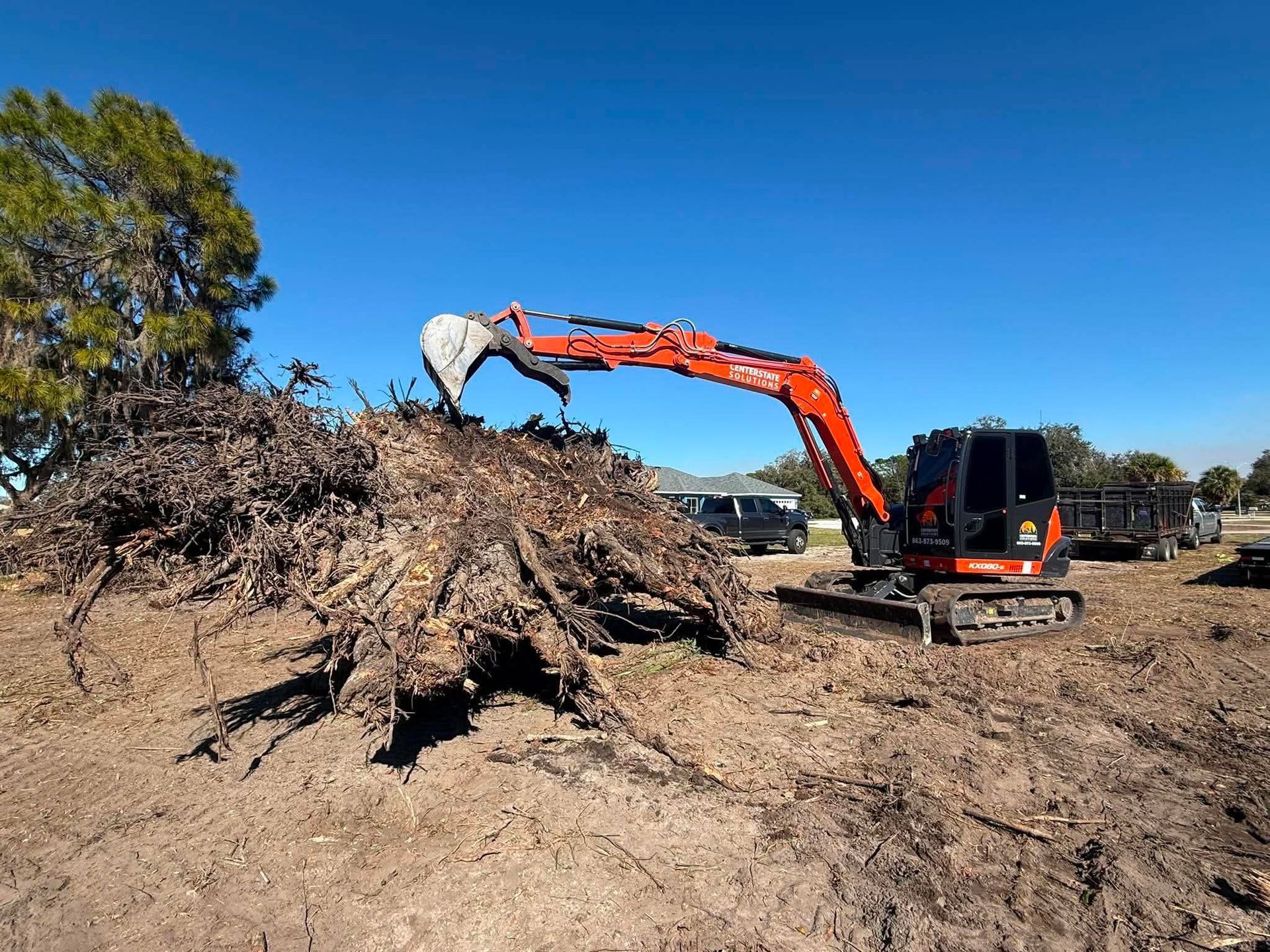 Orange excavator lifting a pile of roots and brush in a dirt lot under a clear blue sky
