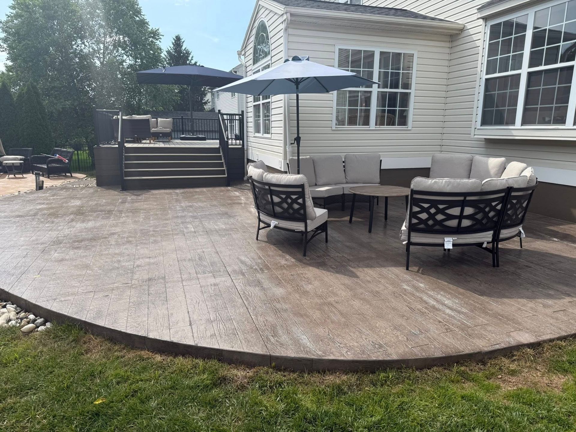 Outdoor patio with concrete surface and seating area, includes a deck, umbrellas, and a house in the background.