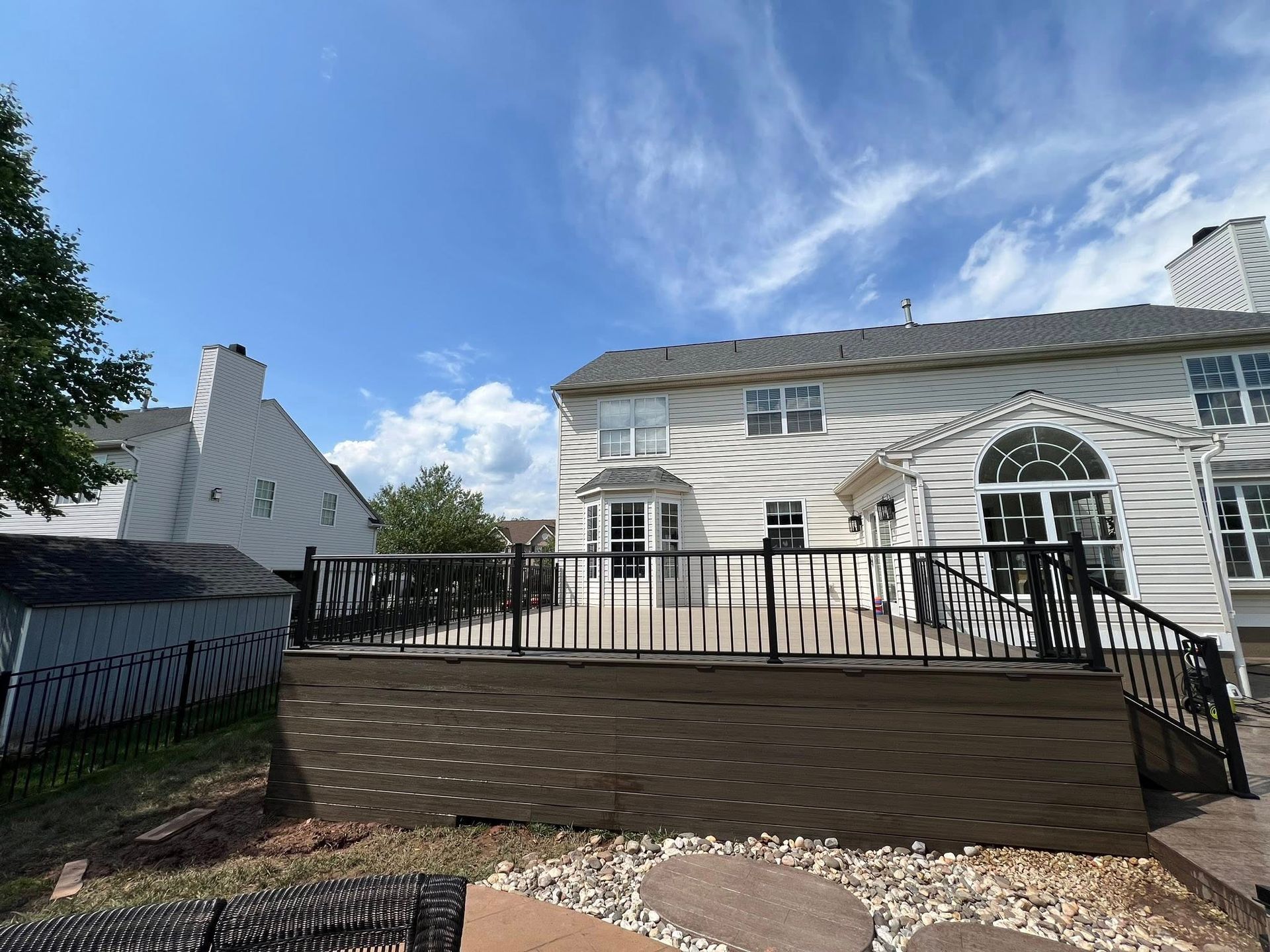 Backyard with a wooden deck, a large house, and a small shed under a bright blue sky with clouds.