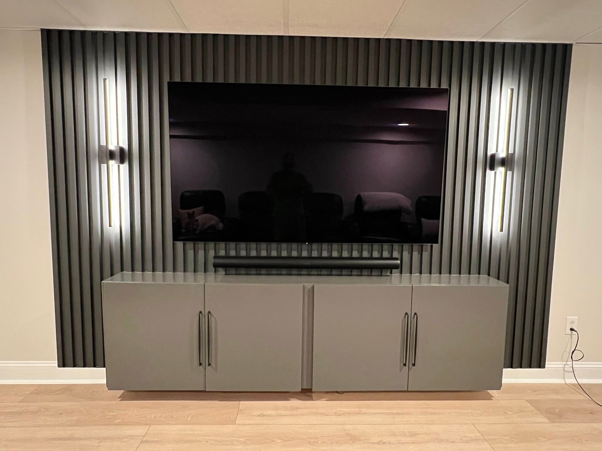 A modern entertainment center featuring a large TV, soundbar, floating grey cabinets, and textured wall with sconce lighting.