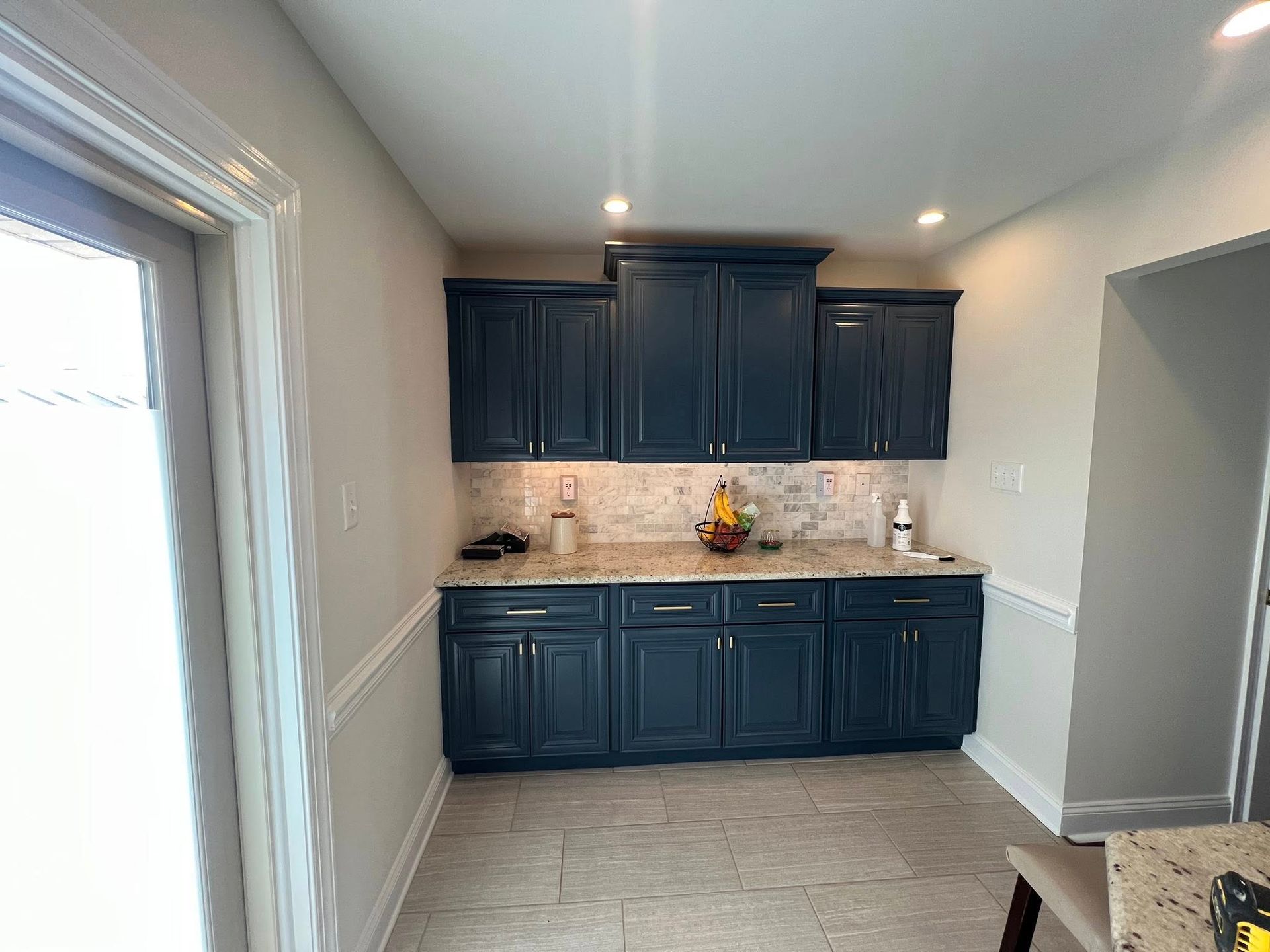 Kitchen with dark blue cabinets, light granite countertops, and beige tile floor. White wainscoting and a doorway on the left.