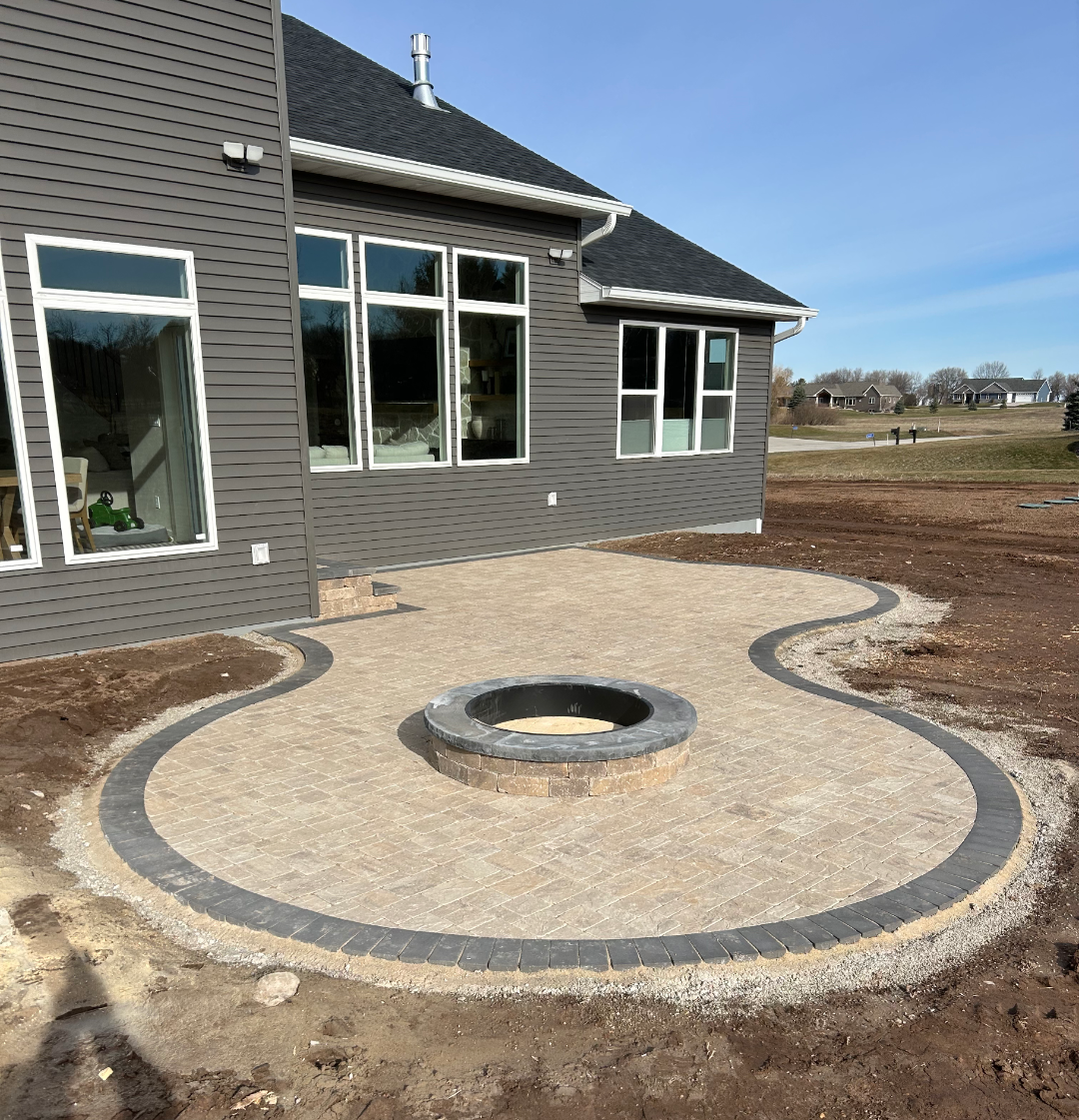 Backyard patio with fire pit, bordered by stone and grey pavers, next to a modern house.