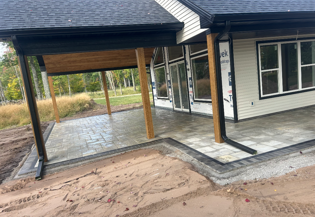 Patio with paver flooring, porch roof, wooden support beams, and a house with white siding and black trim.