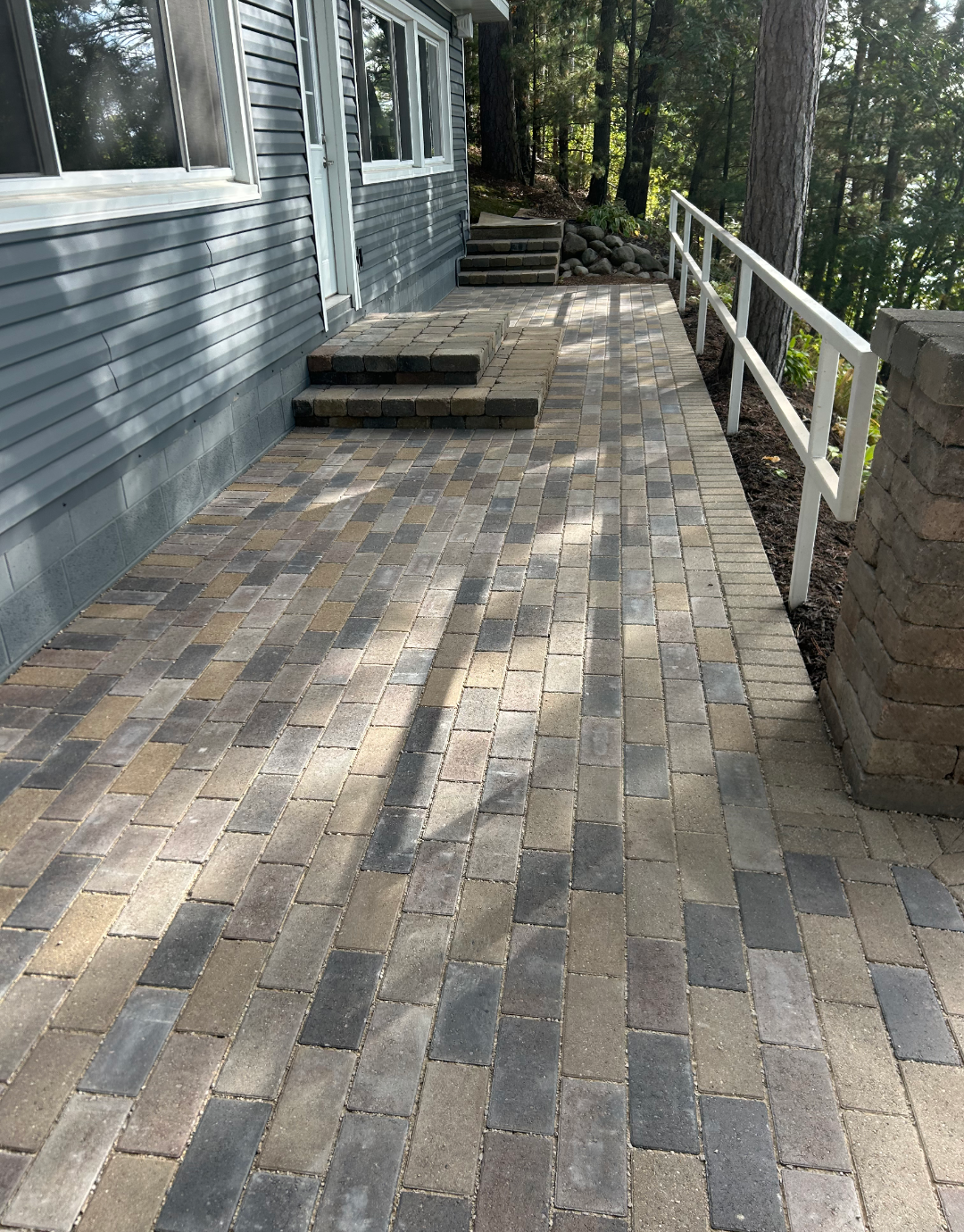 Brick walkway leading to a house with steps and a white railing, surrounded by trees.