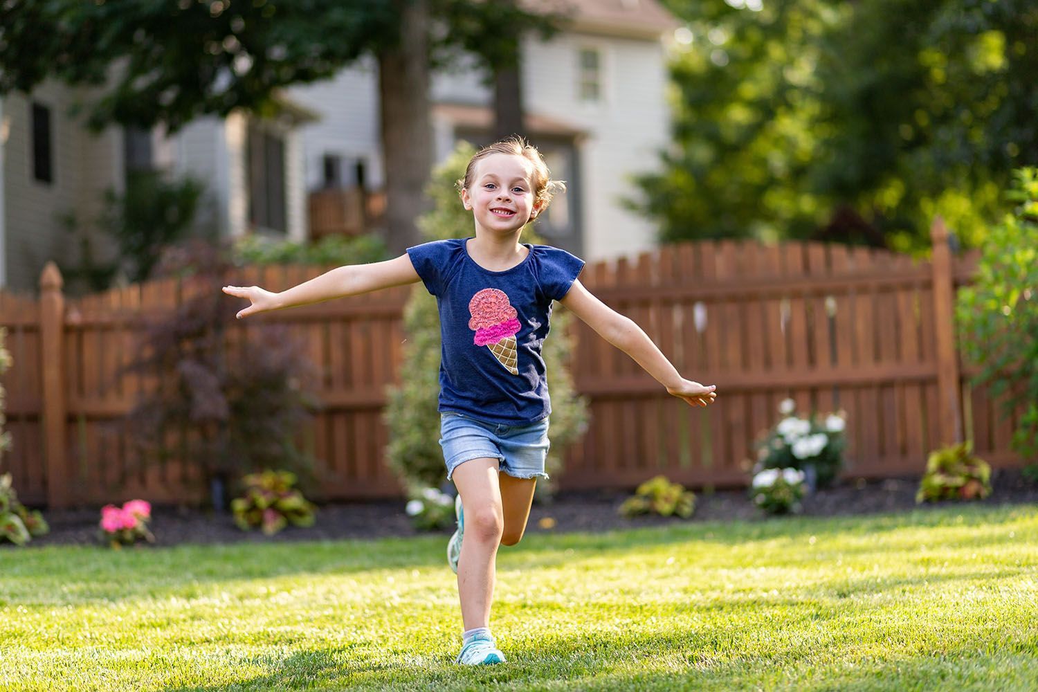 Child running across a sunny backyard with arms outstretched, wearing a blue shirt and shorts.