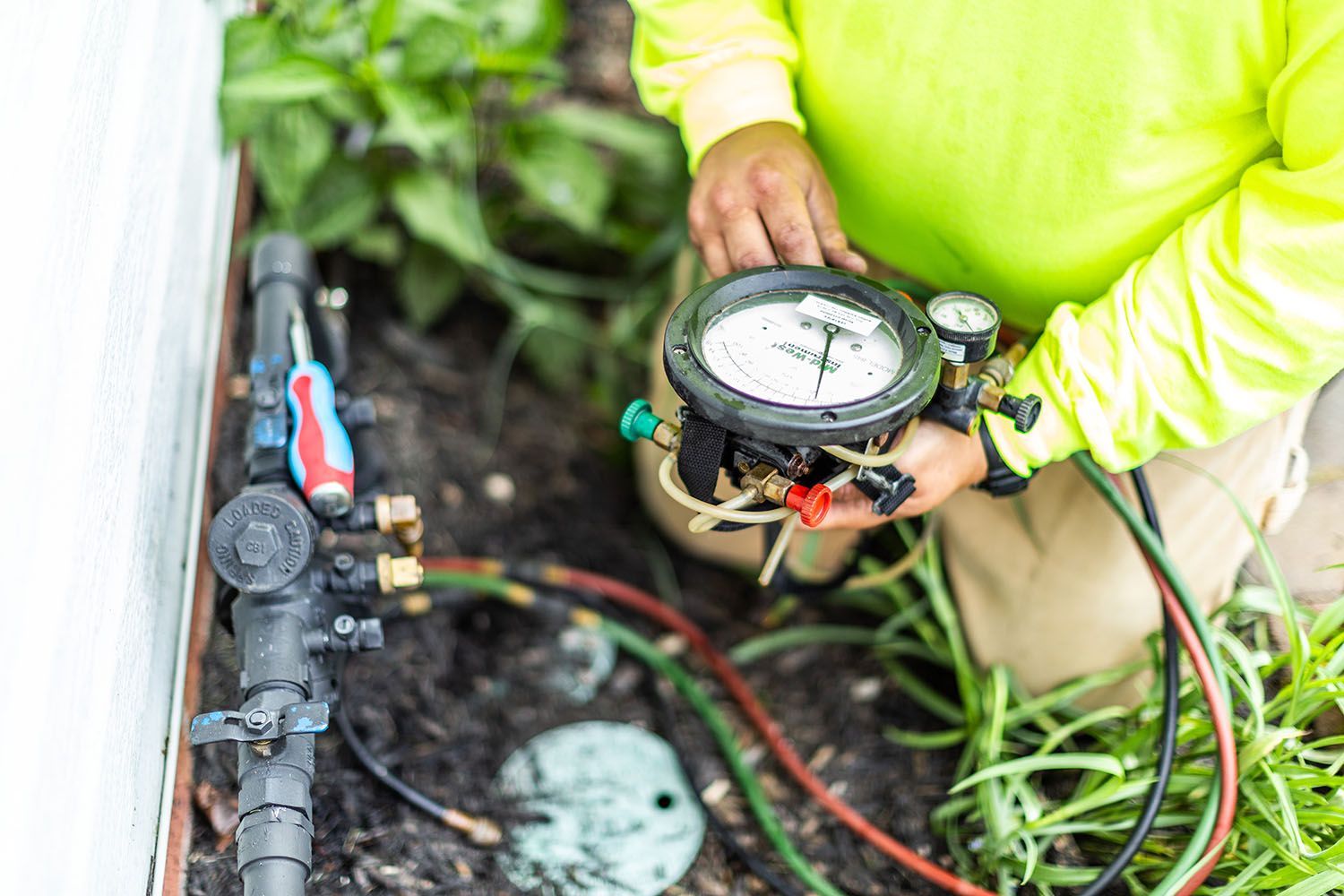 Technician holding a pressure gauge beside outdoor irrigation pipes and valves near a wall