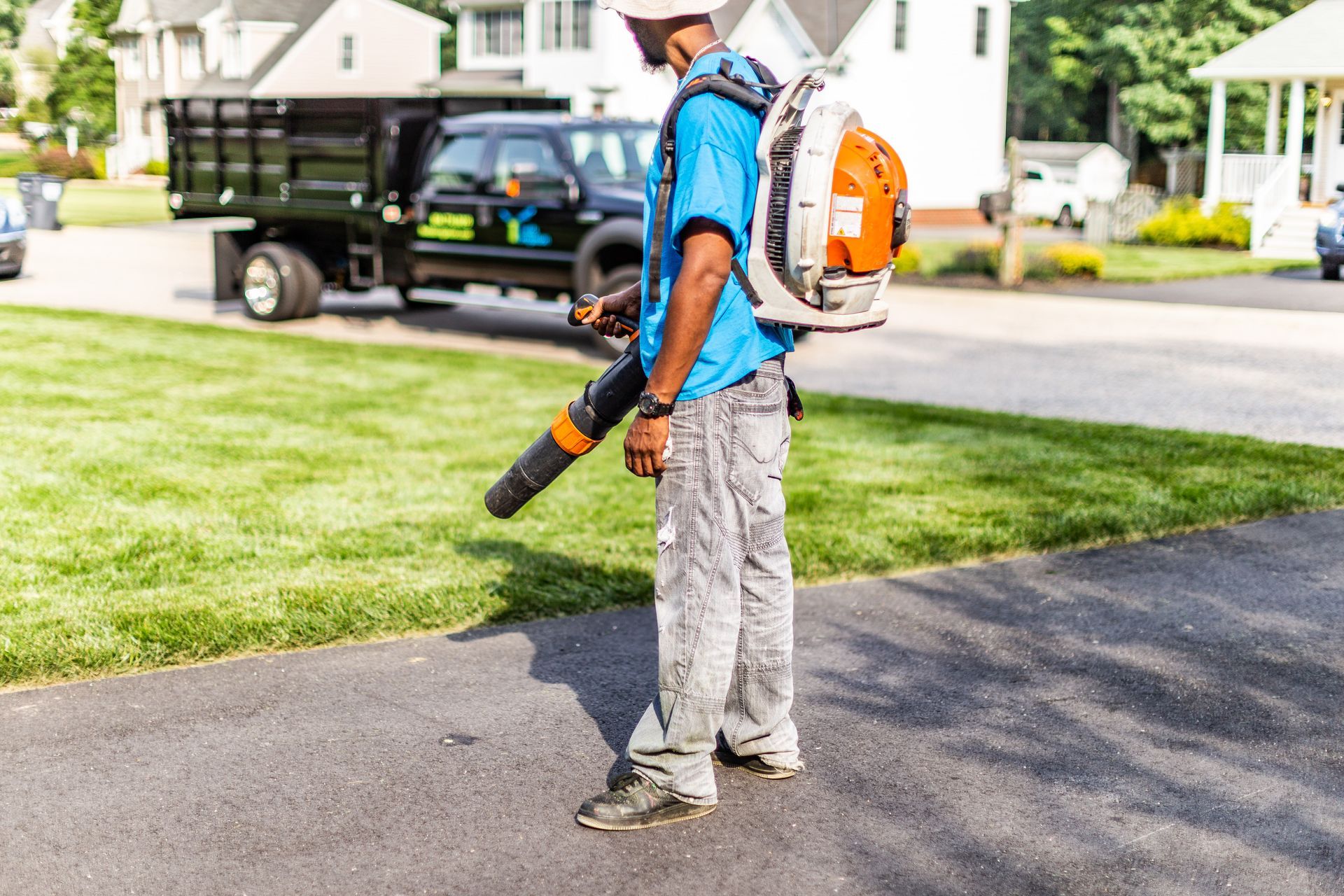 Worker using a leaf blower on a driveway beside a lawn and service truck