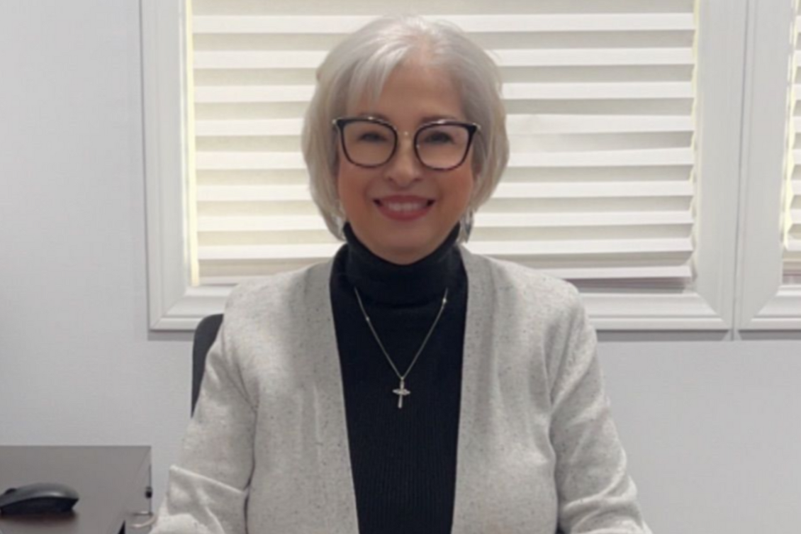 Learn More About Secretarial Services Smiling person with white hair and glasses, wearing a black turtleneck and gray cardigan, seated indoors.