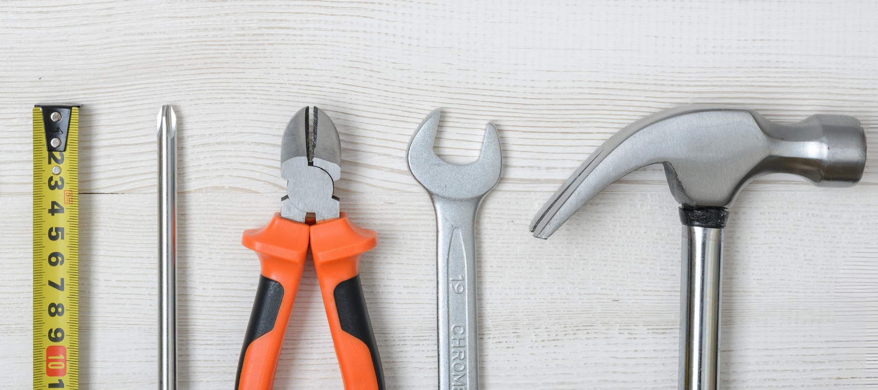 Tools arranged on a white wooden surface. Includes a tape measure, screwdriver, pliers, wrench, and hammer.