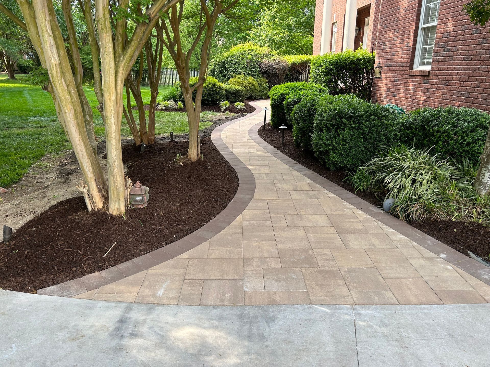 a brick walkway leading to a brick house surrounded by trees and bushes