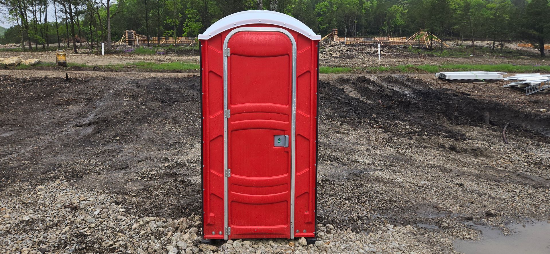A red portable toilet is sitting in the middle of a dirt field.