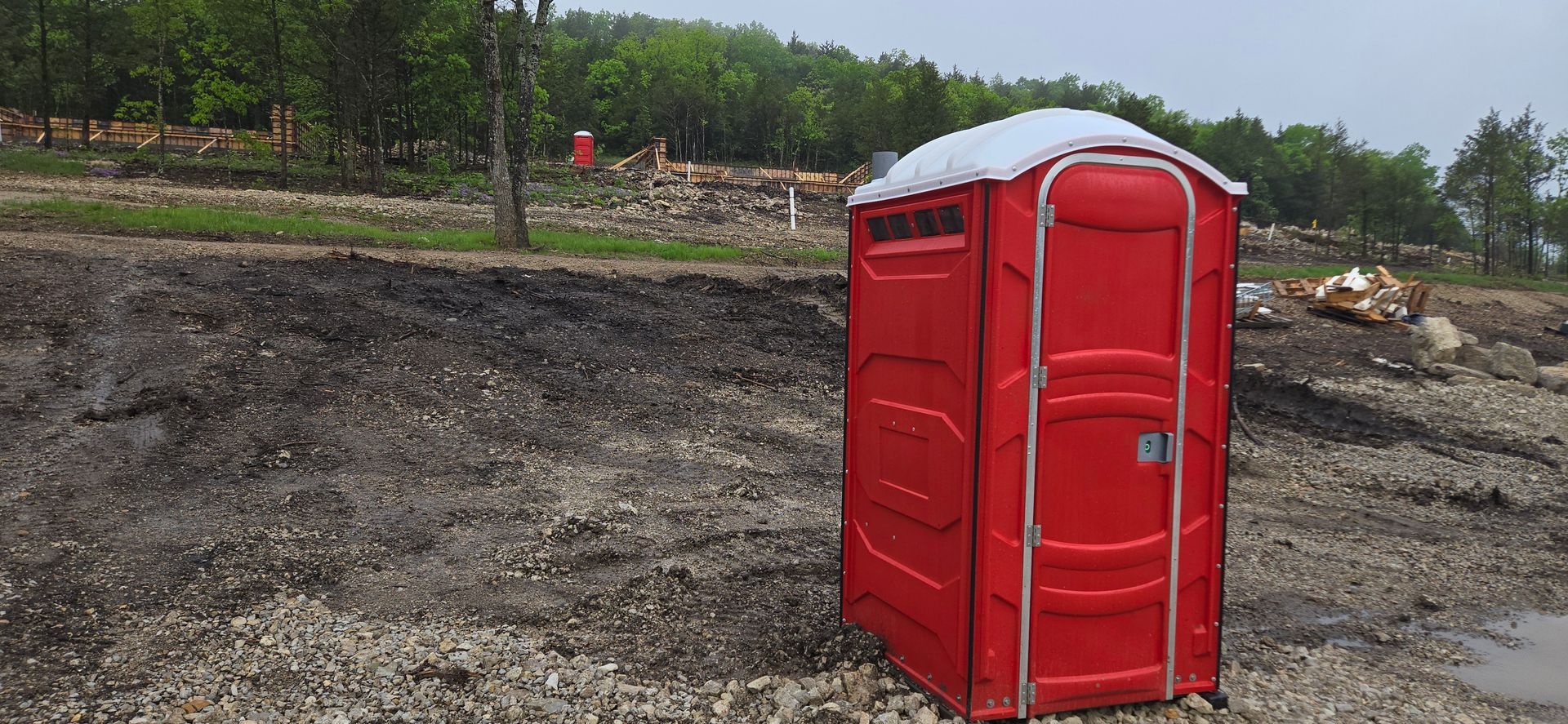 A red portable toilet is sitting in the middle of a dirt field.