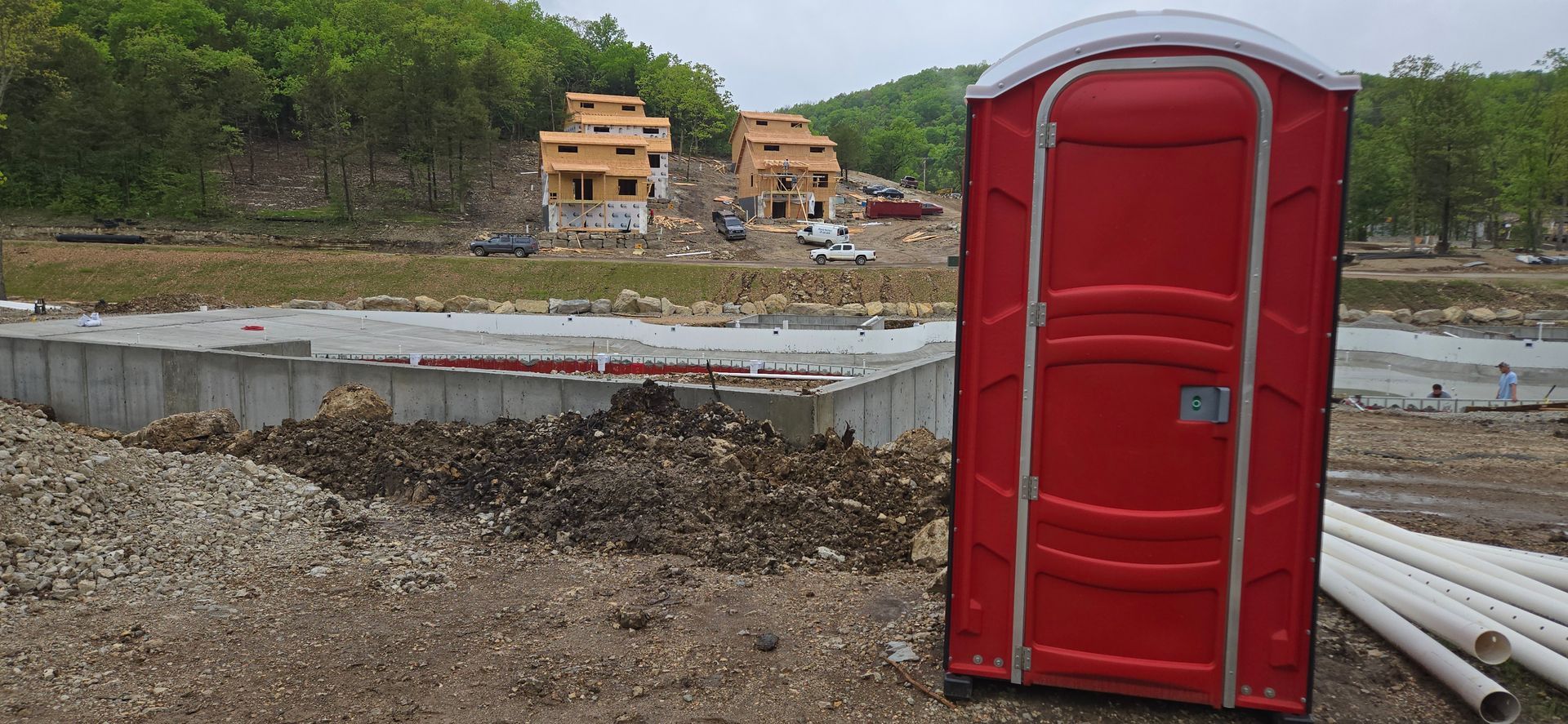 A red portable toilet is sitting in the dirt in front of a construction site.