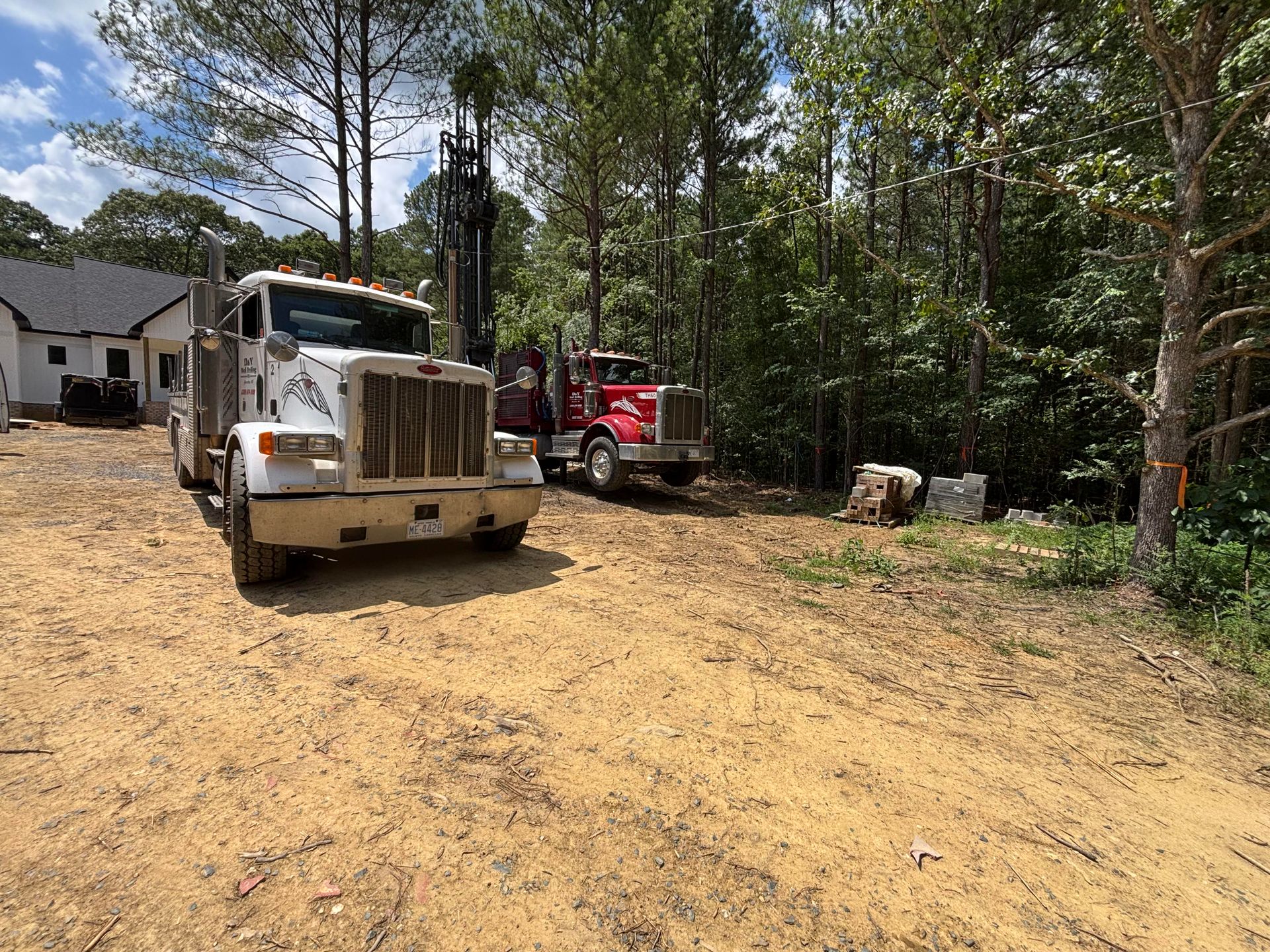 Two semi trucks are parked in a dirt lot in front of a house.