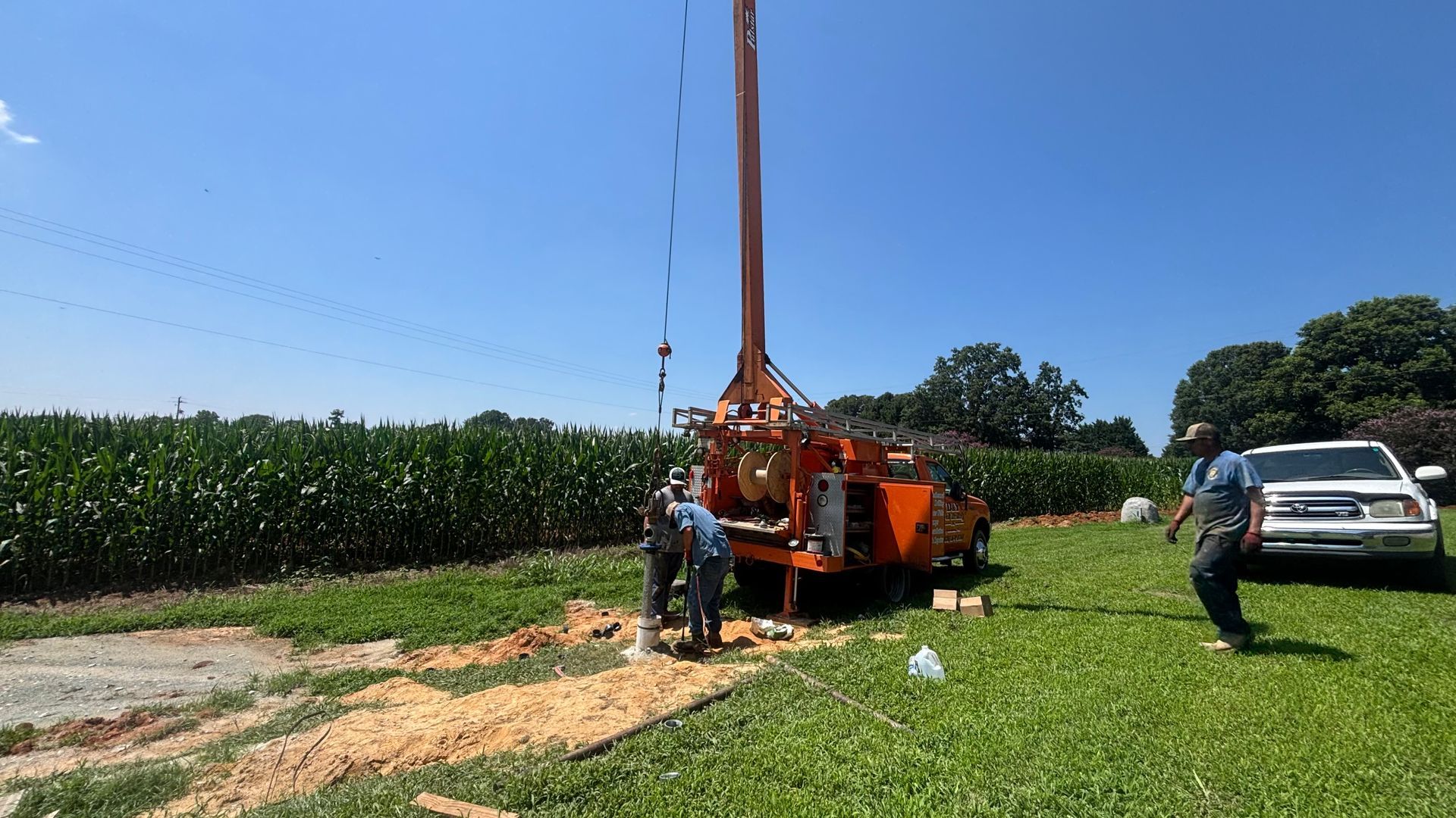 A drilling rig in a field for a well. Two workers attend the orange machine under a bright blue sky.