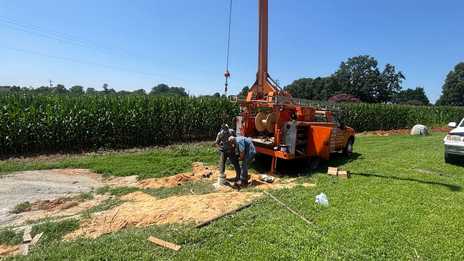 Two workers operating an orange drilling rig in a field next to a cornfield on a sunny day.