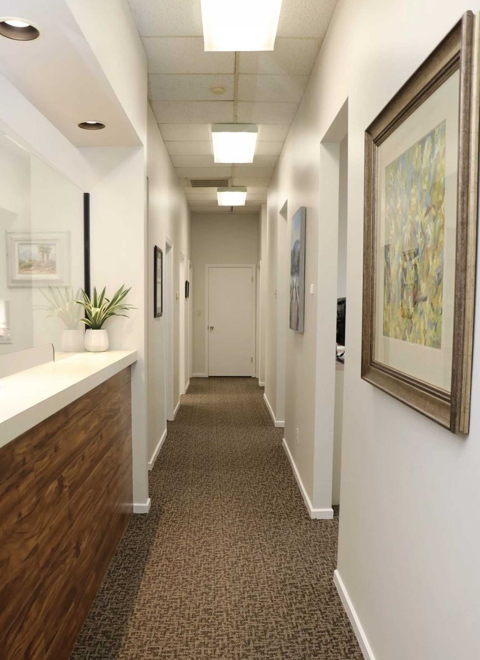 A bright hallway with a wooden reception desk on the left, patterned carpet, and framed art on the white walls.