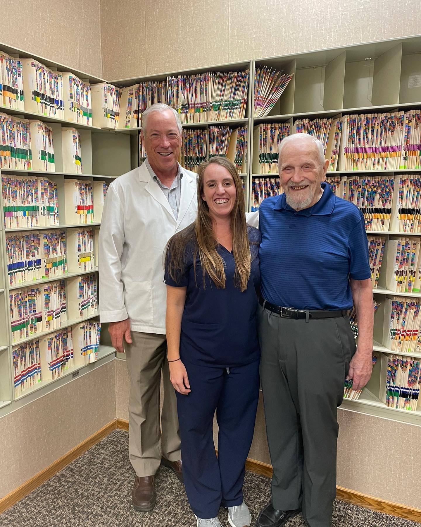 A professional office setting with two staff members in medical attire standing on either side of a man in front of files.