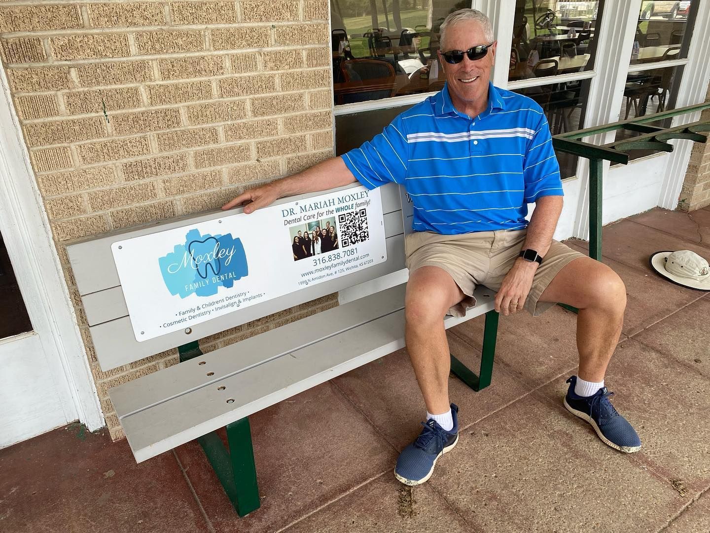 A person wearing sunglasses and a blue striped shirt sits on a bench with a sign against a brick wall.