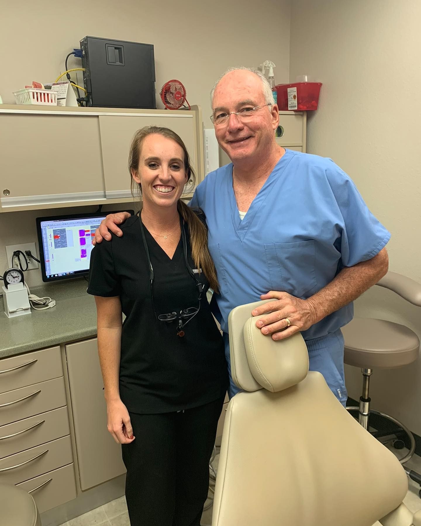 A staff member in black scrubs and a professional in blue scrubs smile together in a dental office.