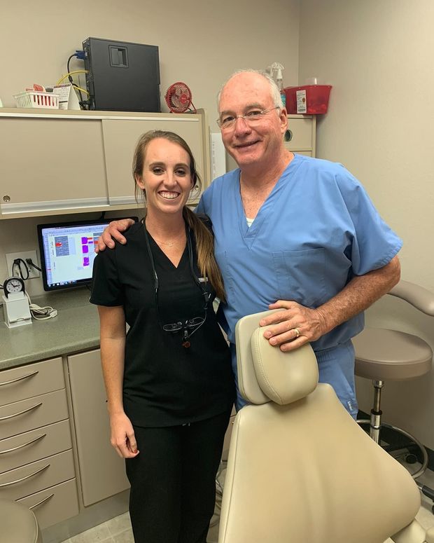 A smiling woman in black scrubs and a man in blue scrubs pose together in a dental office next to a patient chair.