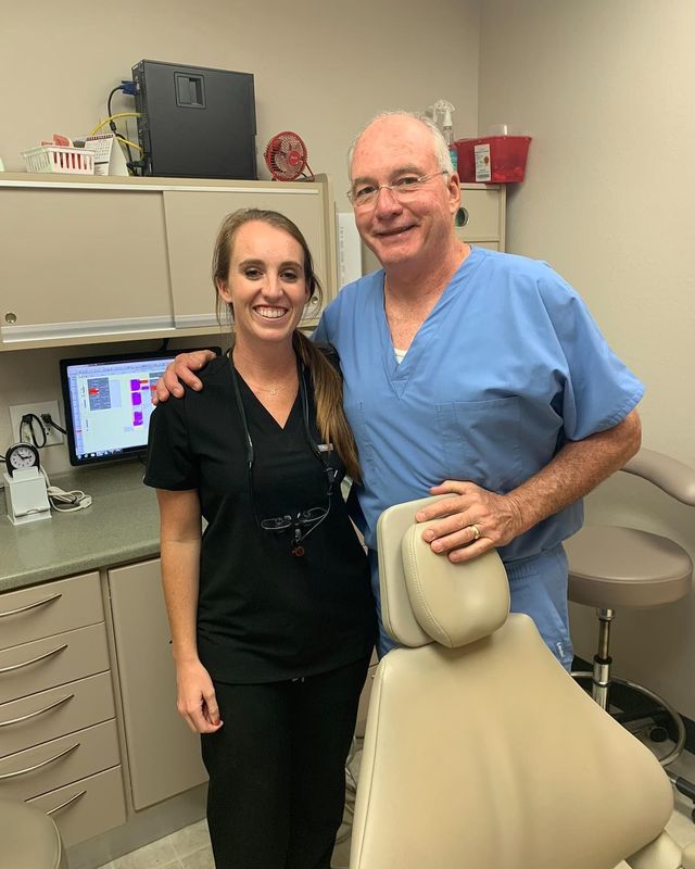 A smiling woman in black scrubs and a man in blue scrubs pose together in a dental office next to a patient chair.